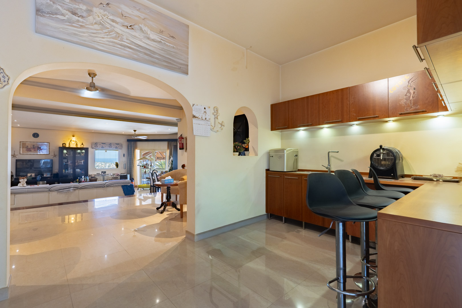Bar intégré avec mobilier en bois, tabourets hauts et éclairage sous les placards, s'ouvrant sur le salon par une arche décorative et un sol en grès cérame.