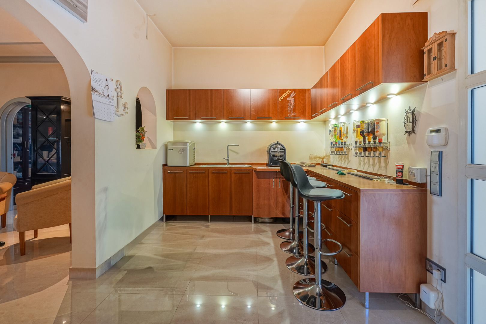 Bar intérieur avec meubles en bois, évier intégré et distributeurs muraux. Comprend des tabourets chromés, un éclairage LED sous les placards et un sol en grès poli.