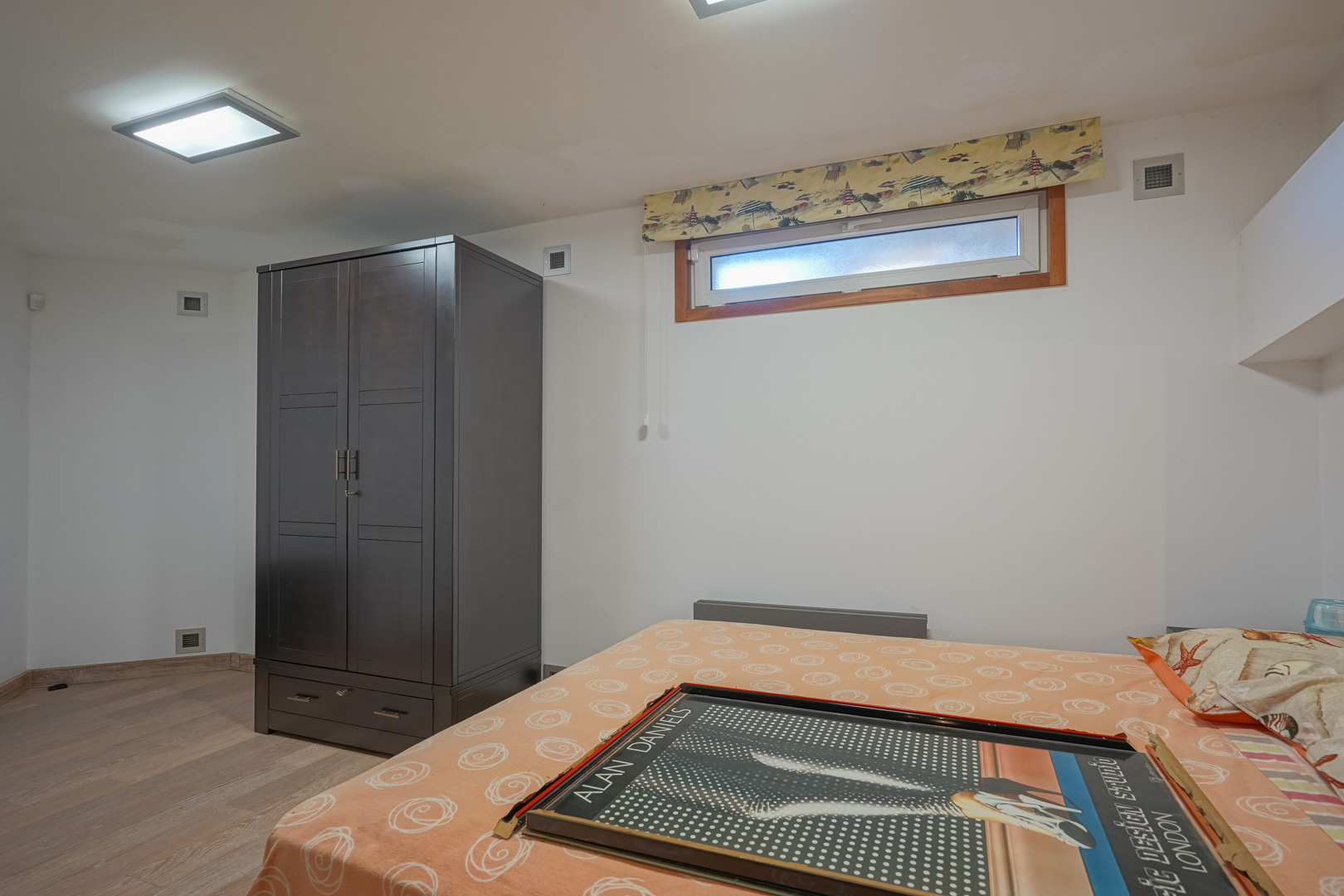 Chambre fonctionnelle avec une armoire en bois sombre, un sol stratifié clair et une fenêtre horizontale sous un éclairage de plafond encastré.
