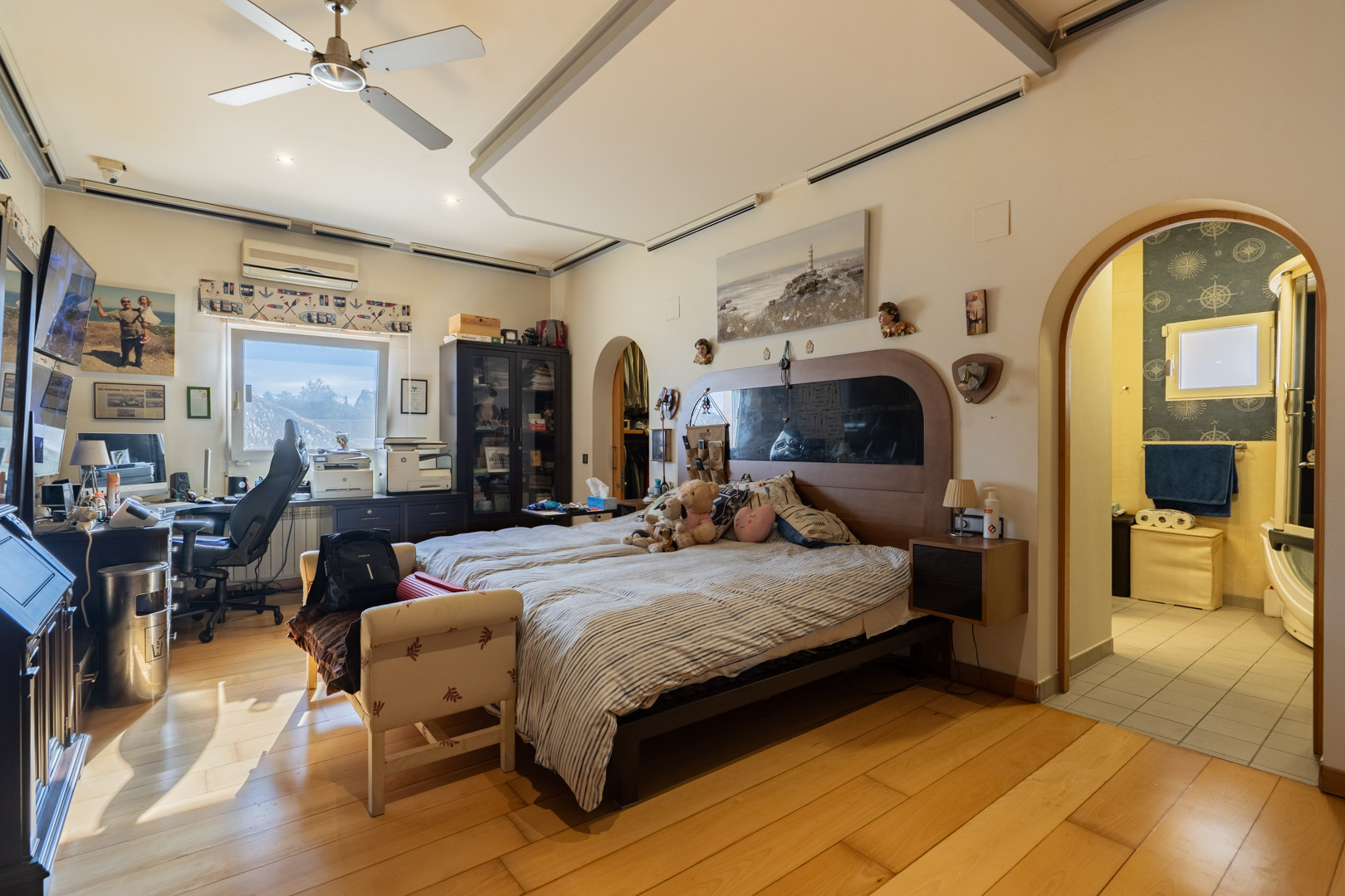 Chambre principale avec parquet en bois noble, espace bureau intégré et salle de bain attenante. La pièce dispose d'un ventilateur de plafond et d'une climatisation.