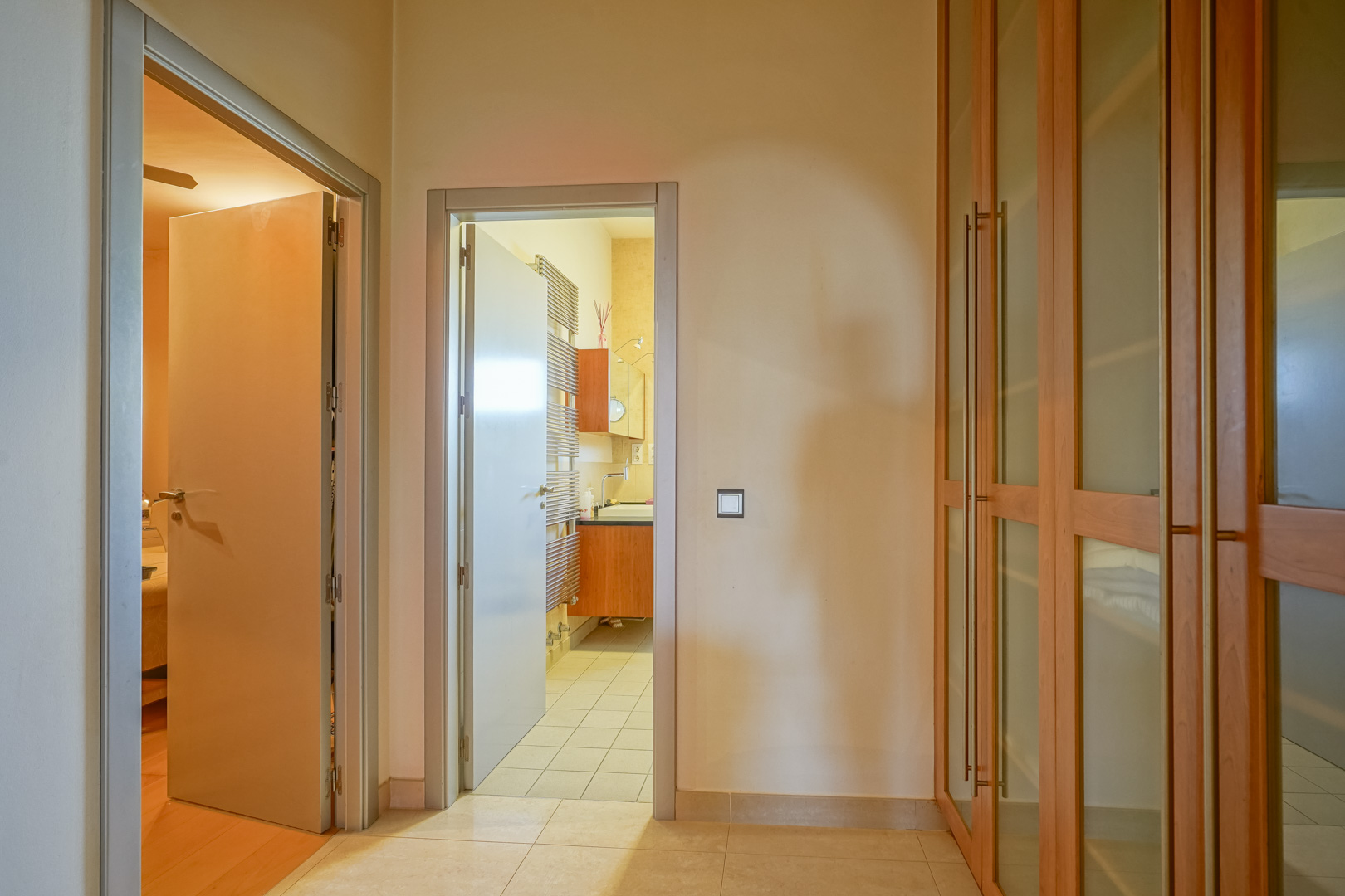 Couloir de distribution avec placards intégrés en bois et verre, reliant une chambre au sol en bois noble à une salle de bains attenante avec radiateur sèche-serviettes.