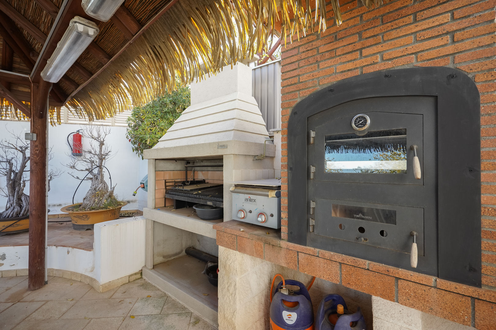 Cuisine d'été équipée d'un four Tromen encastré, d'un barbecue et d'une plancha, le tout abrité par une pergola en bois au toit de chaume.