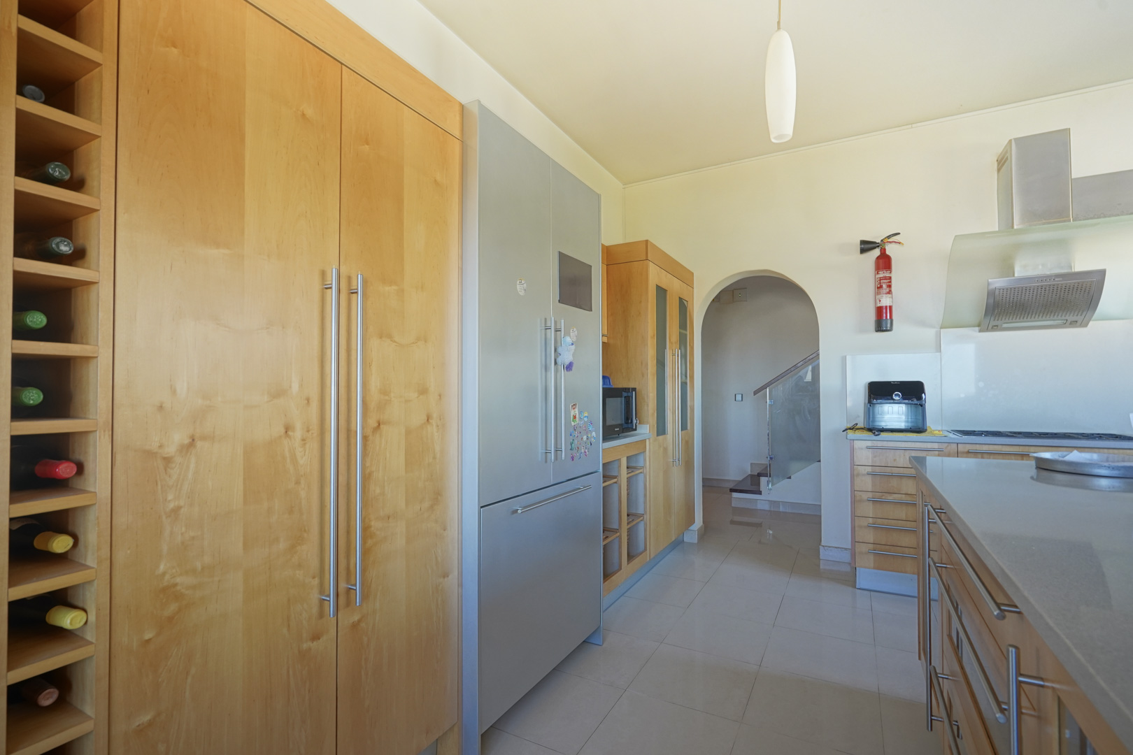 Cuisine équipée de meubles en bois sur mesure, d'un casier à vin intégré et d'appareils en inox. L'arche s'ouvre sur un escalier doté d'une rampe en verre contemporaine.