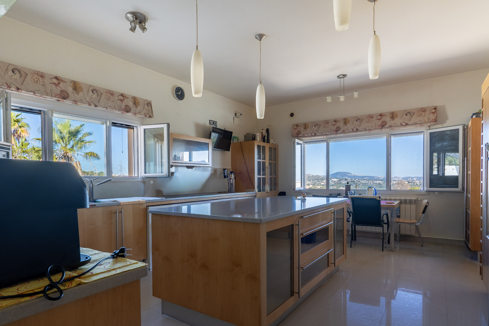 Cuisine spacieuse avec îlot central en bois et plans de travail gris. Les grandes fenêtres offrent une vue panoramique sur la mer et les montagnes environnantes.