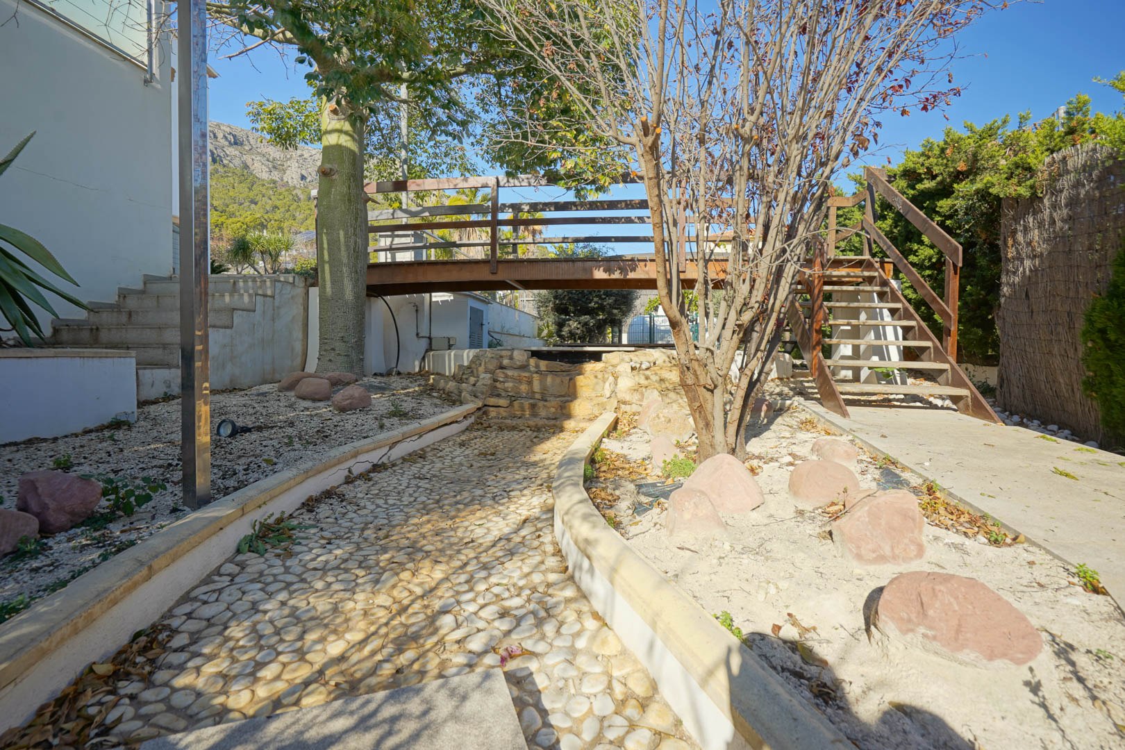 Espace jardin avec passerelle en bois sur une cascade décorative en pierre, chemin pavé de galets et escaliers, offrant une vue sur la Sierra de Oltamar.