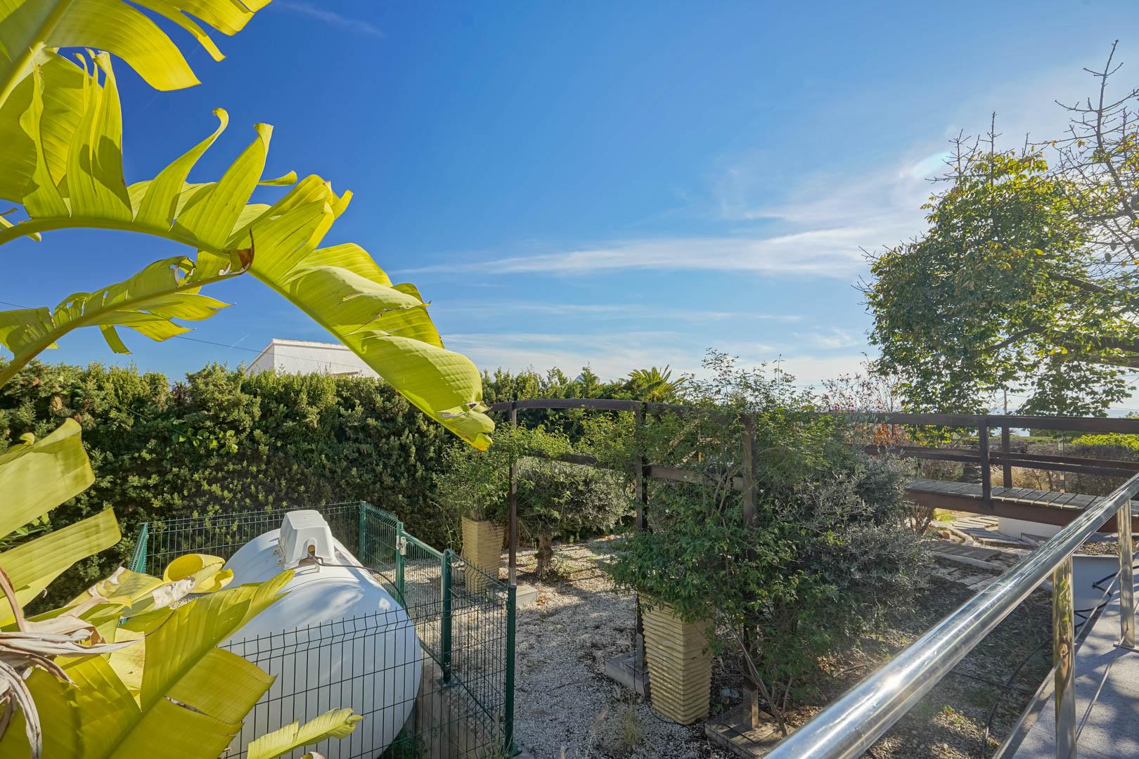 Espace jardin latéral avec réservoir utilitaire clôturé, passerelle en bois et feuillage tropical sous un ciel bleu.