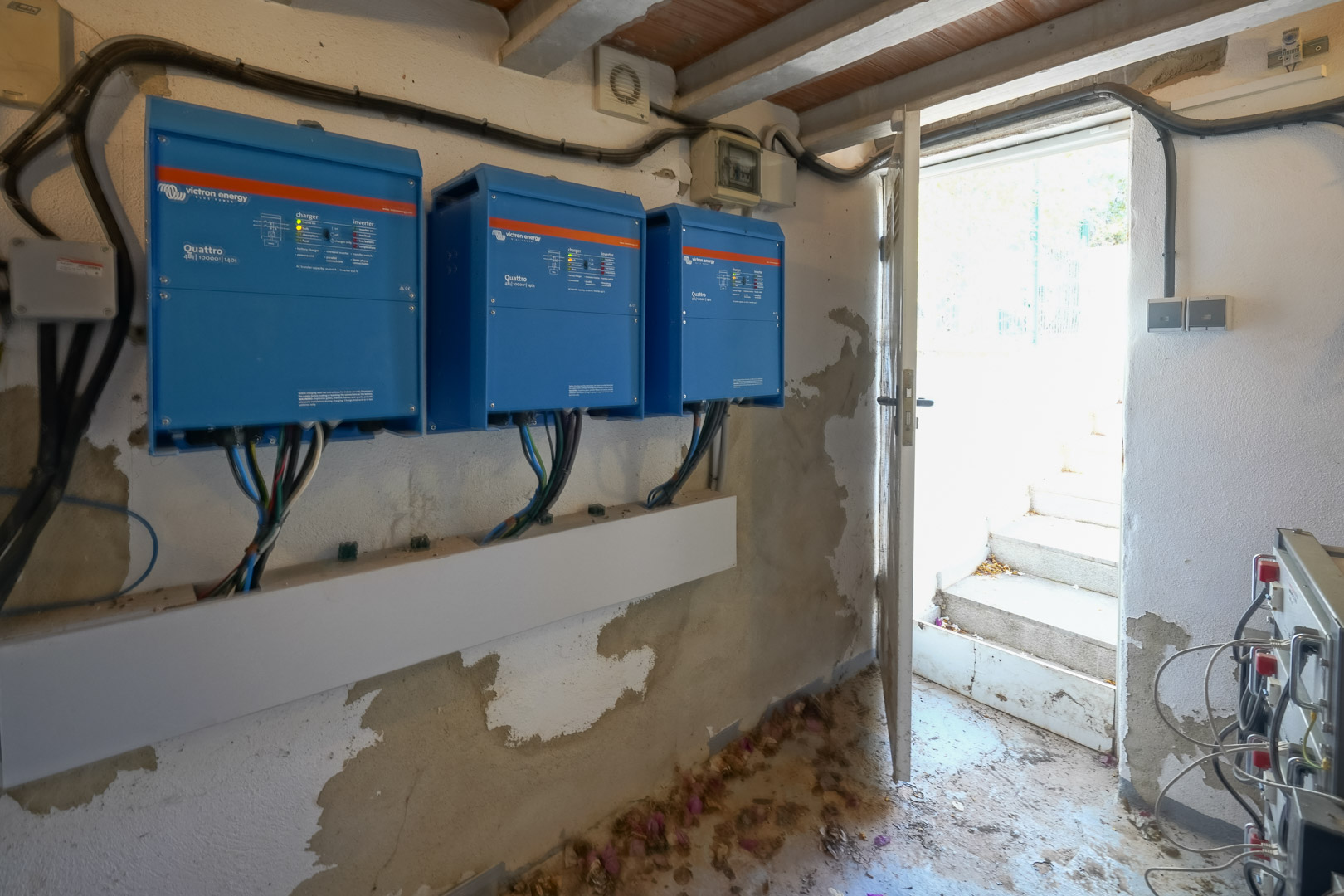 Local technique équipé de trois onduleurs Victron Energy Quattro pour l'installation solaire, situé près d'une sortie vers l'extérieur.