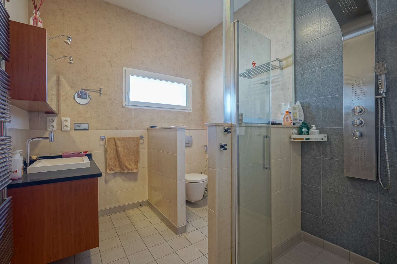 Salle de bain avec colonne de douche hydromassante, paroi en verre et toilettes séparées. Équipée d'un meuble en bois, plan de travail sombre et papier peint cartographique.