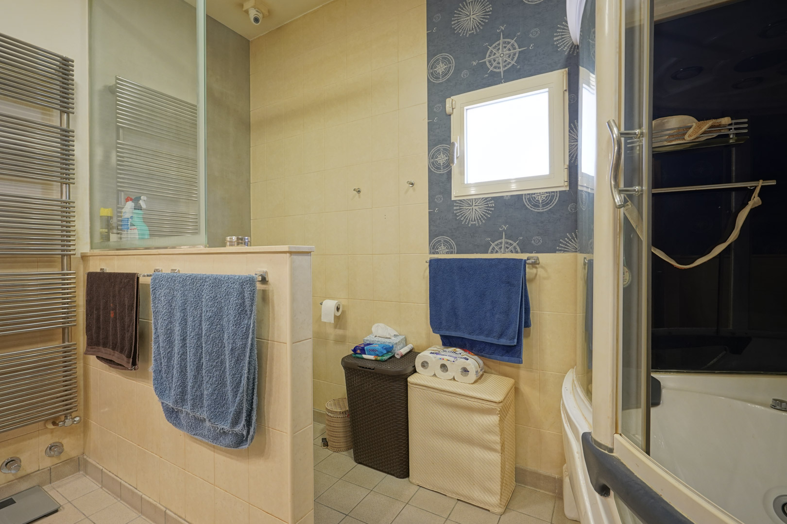Salle de bain fonctionnelle avec carrelage crème, radiateur sèche-serviettes en acier et coin toilette séparé. Un papier peint à motif nautique encadre la fenêtre.