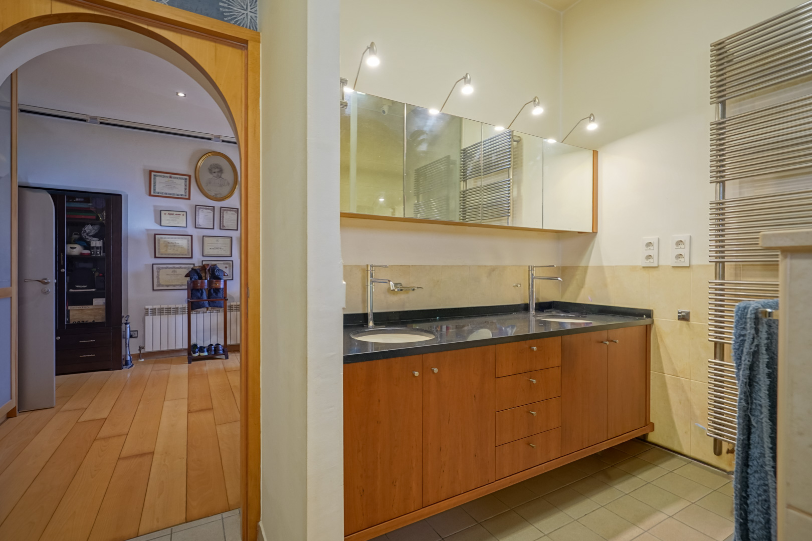 Salle de bains avec double vasque en bois, plan de travail noir et miroirs éclairés. Une arche mène à un dressing avec parquet en bois noble.