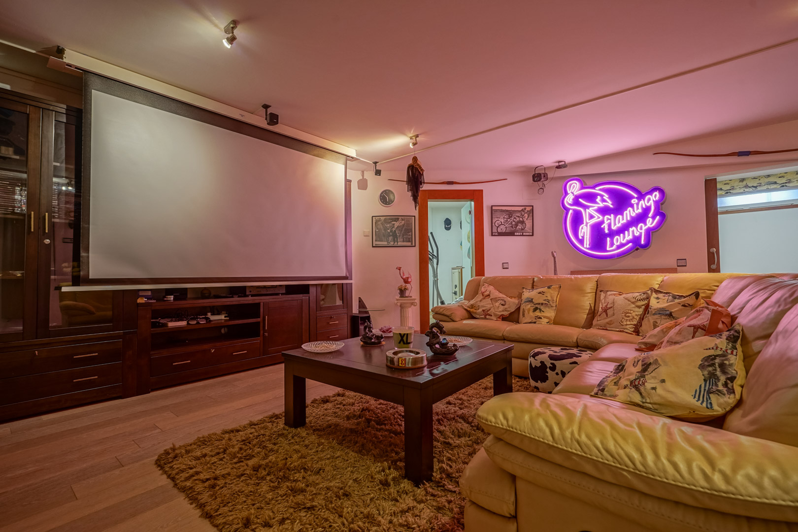 Salle multimédia avec écran de projection, mobilier en bois sombre et canapé d'angle en cuir. L'espace dispose d'un éclairage d'ambiance au néon et d'un sol en bois.
