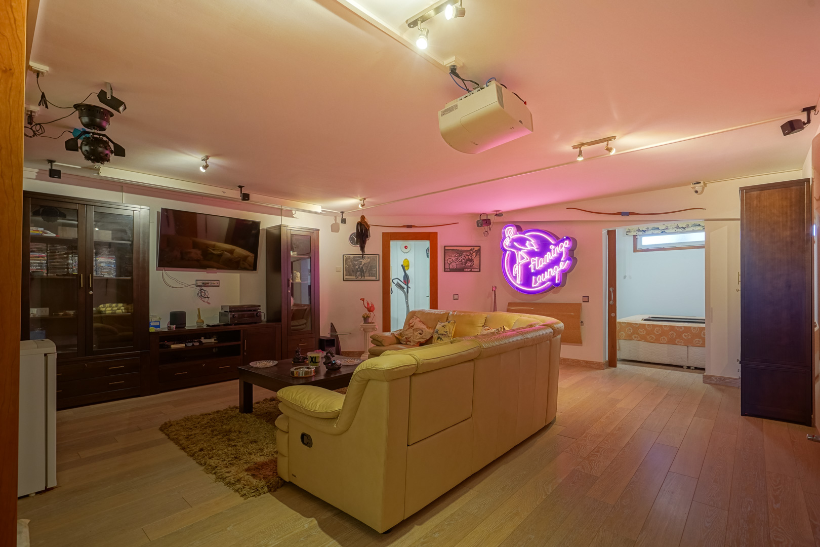 Salle multimédia avec parquet, projecteur au plafond et canapé en cuir. L'espace dispose de meubles sombres, d'un éclairage sur rails et d'une enseigne lumineuse au néon.