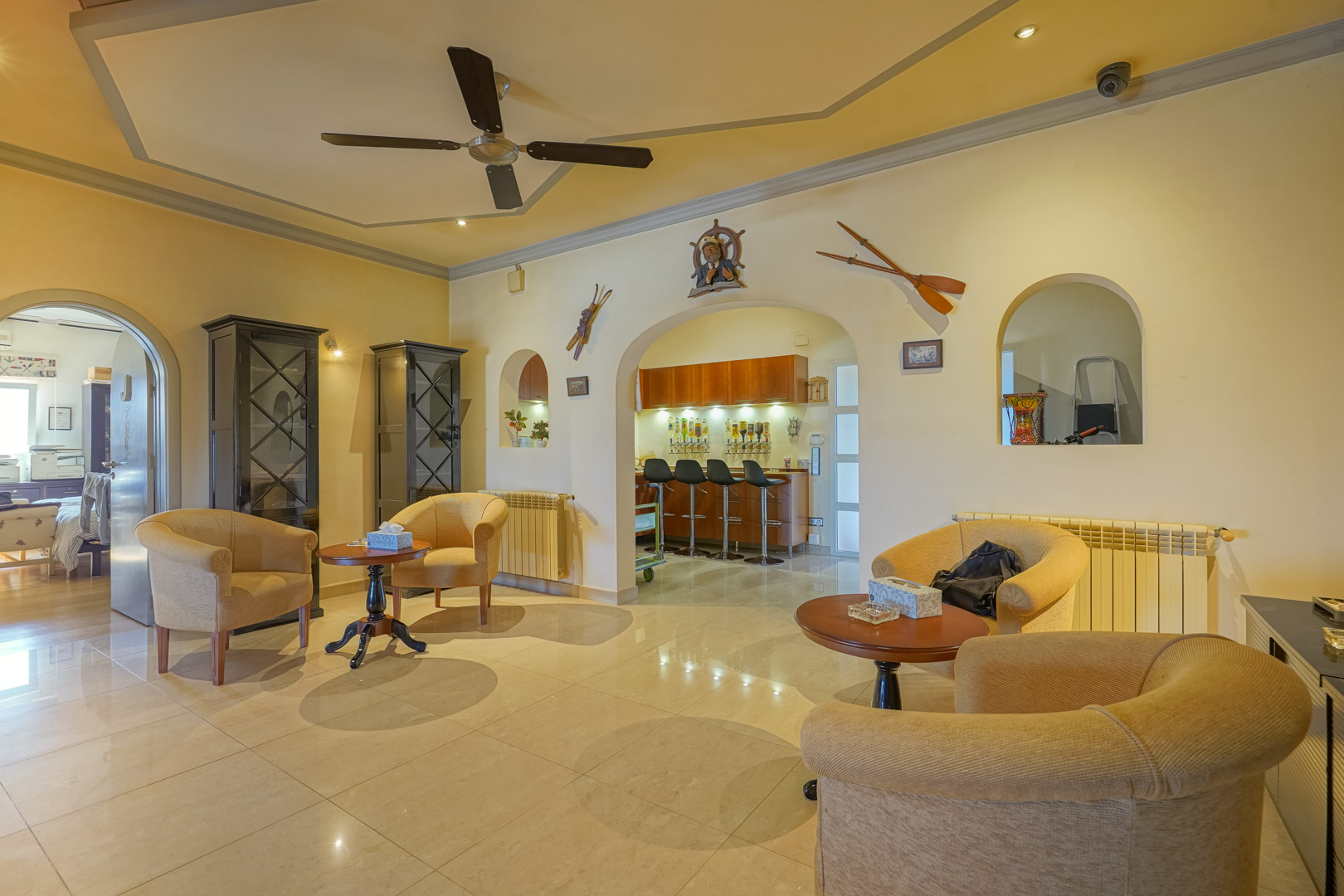 Salon spacieux avec ventilateur de plafond et sol carrelé, incluant un bar intégré accessible par des arches élégantes ornées de motifs nautiques.