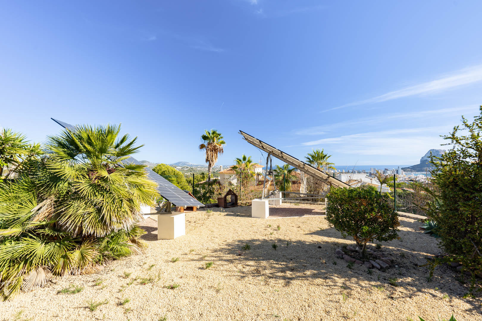 Système de panneaux solaires motorisés sur socles en béton dans un jardin de gravier, avec vue panoramique sur le Peñón de Ifach et Calpe.
