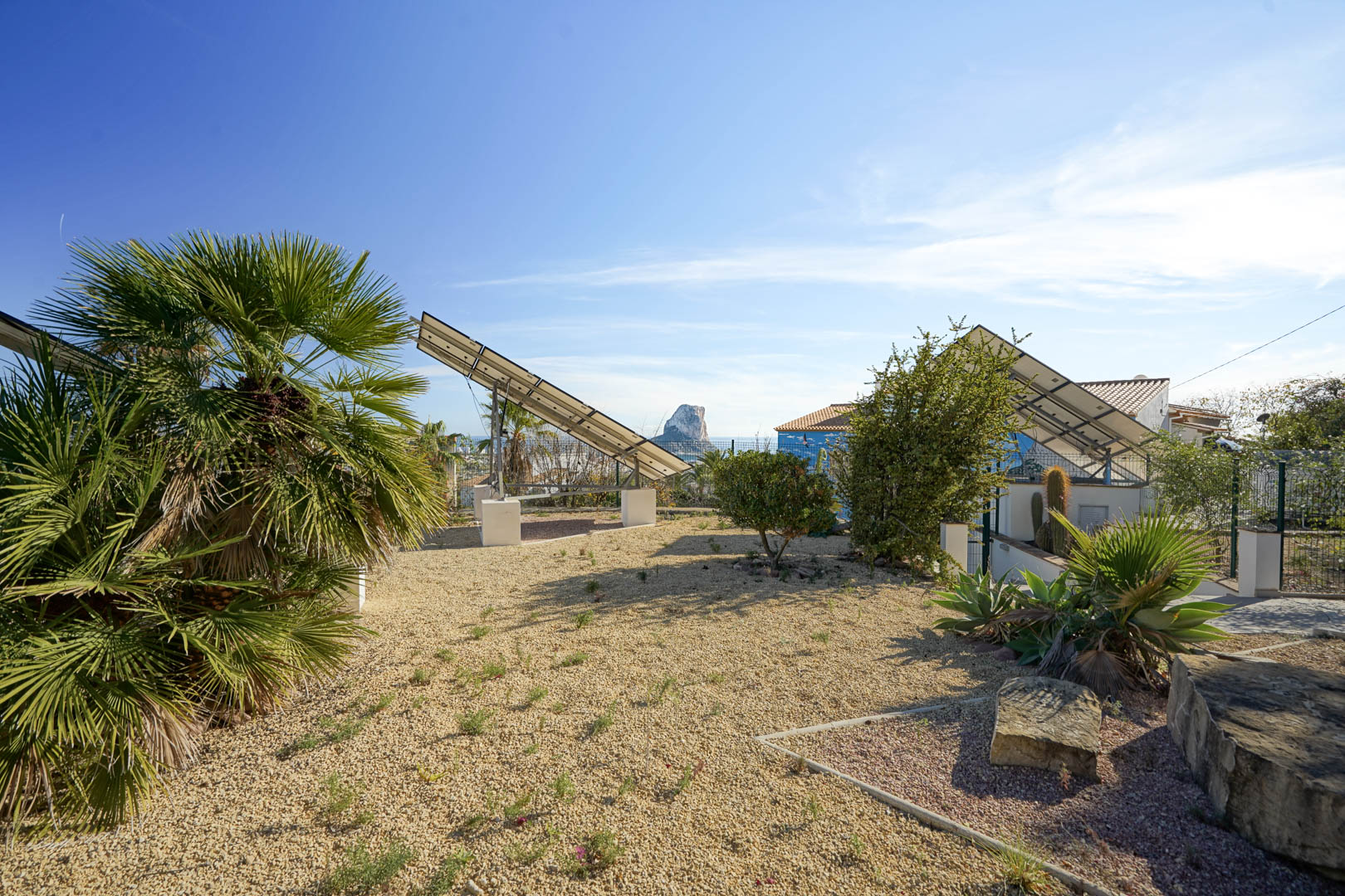 Système photovoltaïque avec tours solaires motorisées dans un jardin de gravier, offrant une vue dégagée sur le Peñón de Ifach et la flore locale.