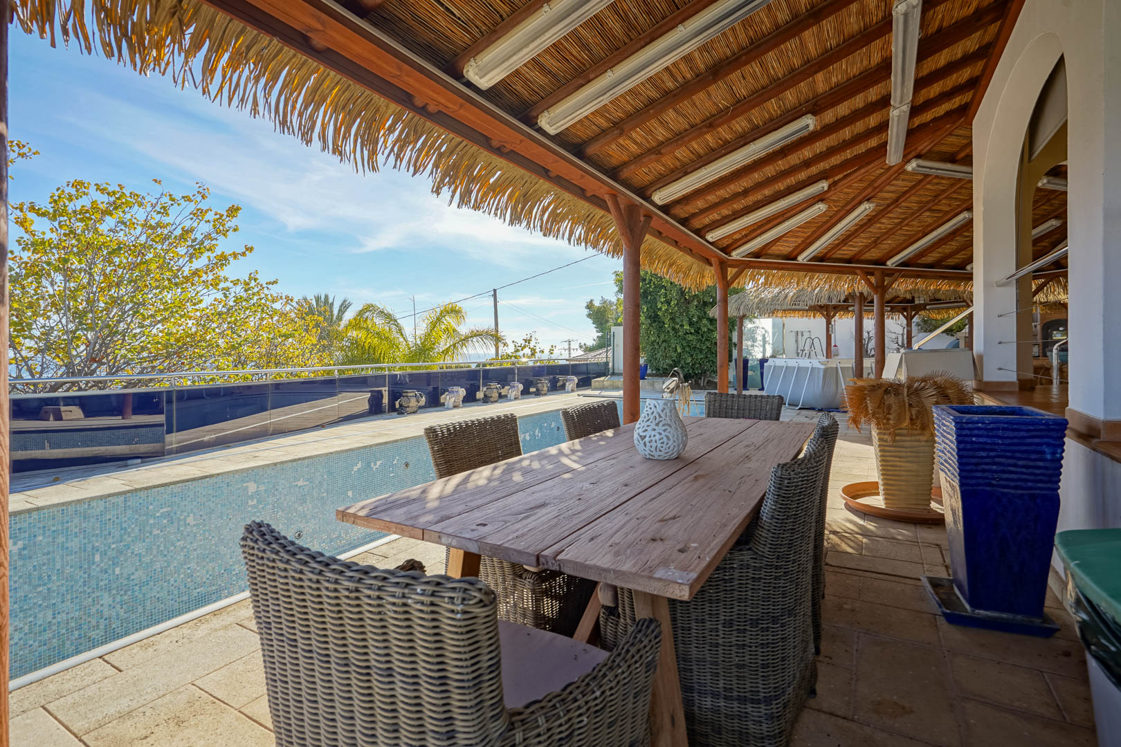 Terrasse couverte avec pergola en chaume, table à manger en bois rustique et chaises en osier, située à côté de la piscine avec vue sur le jardin.