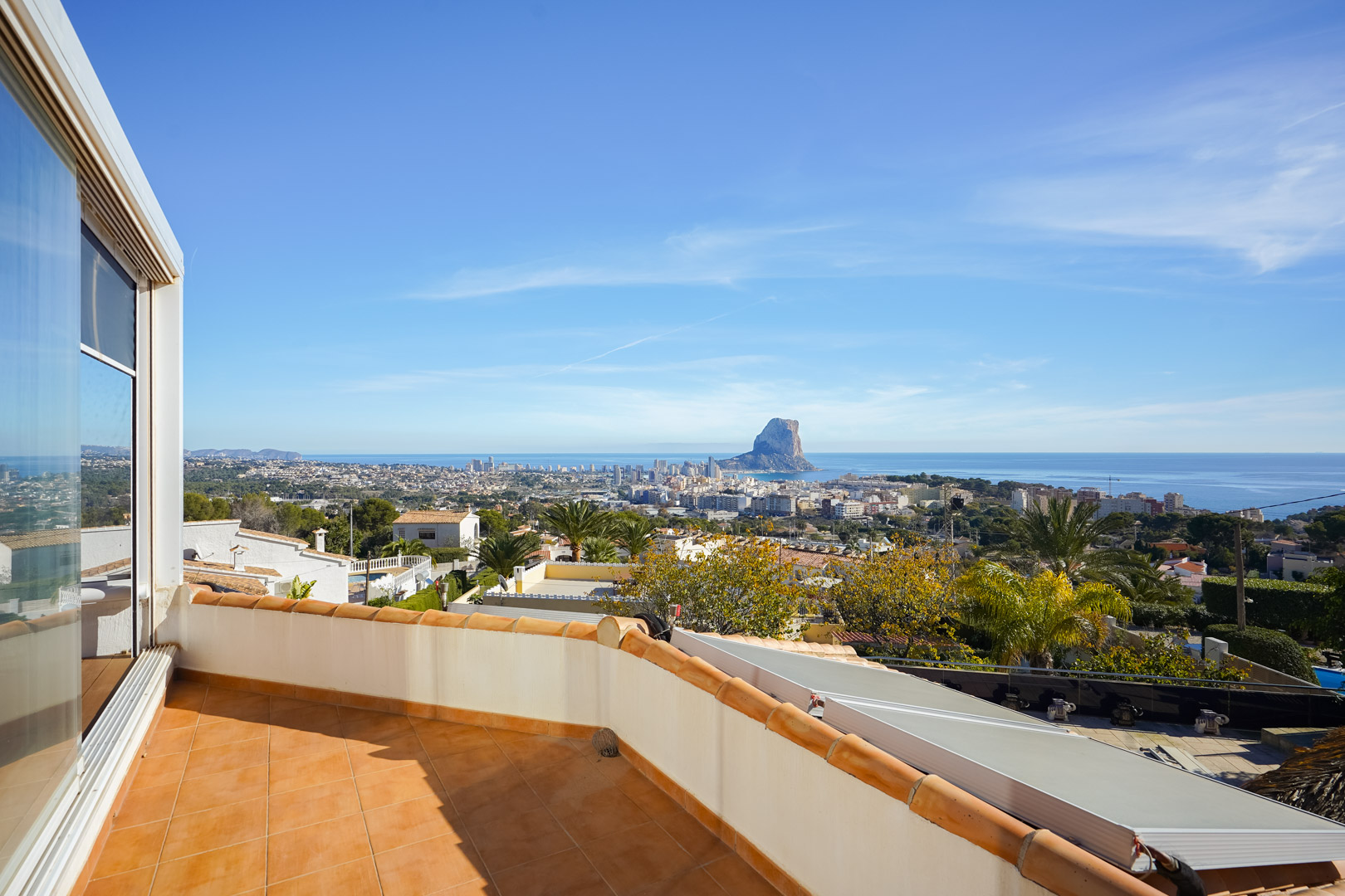 Terrasse supérieure avec carrelage en terre cuite offrant une vue panoramique sur le Peñón de Ifach et Calpe. Des panneaux solaires sont installés sur le toit inférieur.