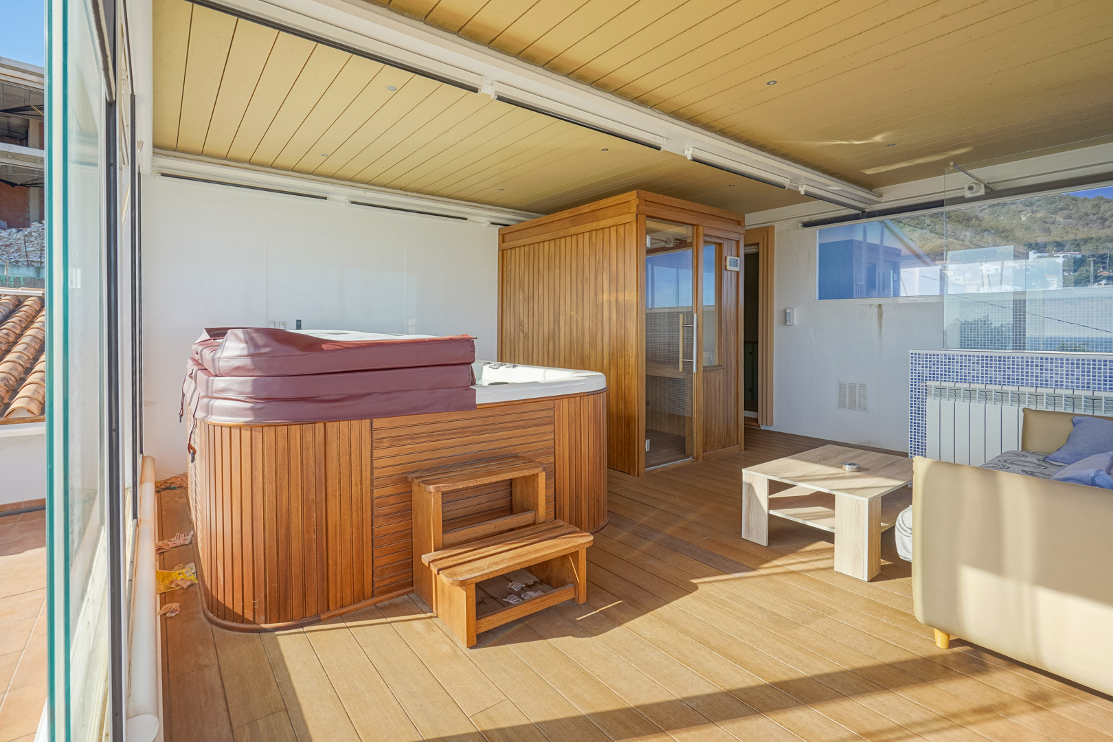 Terrasse supérieure vitrée abritant un espace bien-être avec jacuzzi habillé de bois, sauna privé et sol en parquet, le tout sous un plafond technique avec éclairage intégré.