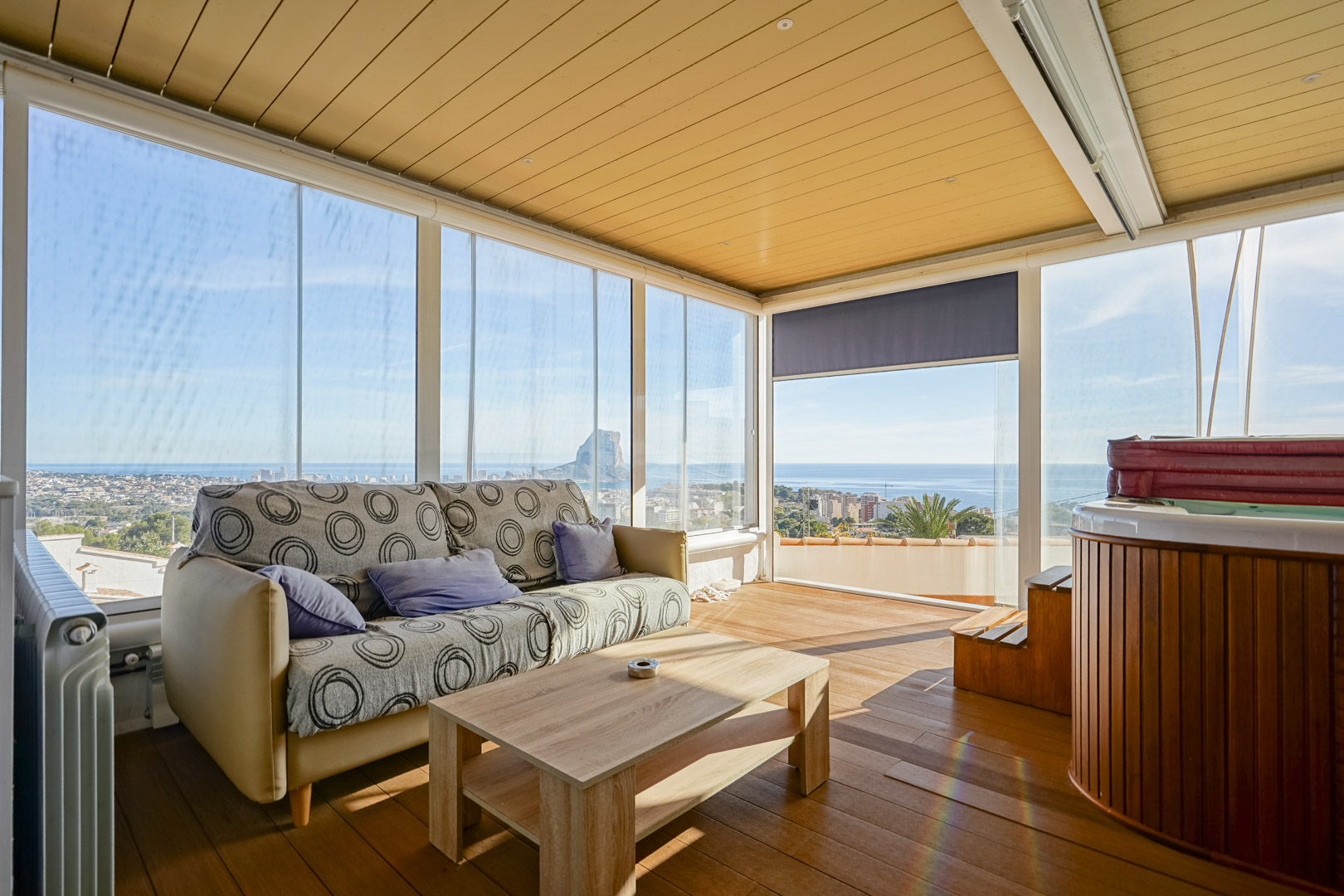 Terrasse supérieure vitrée avec jacuzzi, parquet en bois et vue panoramique sur le Peñón de Ifach. L'espace comprend un coin salon et des parois en verre.