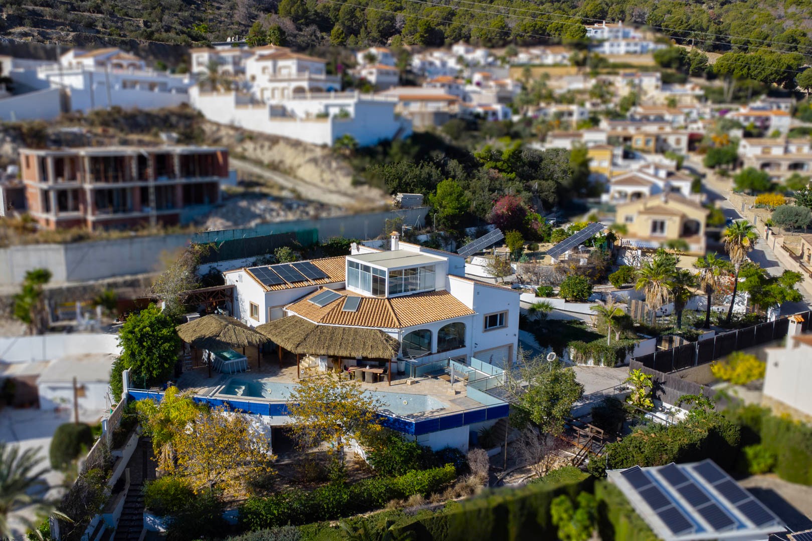 Vue aérienne de la villa illustrant l'espace piscine avec pergolas en chaume, les panneaux solaires en toiture et les suiveurs solaires motorisés dans le jardin.