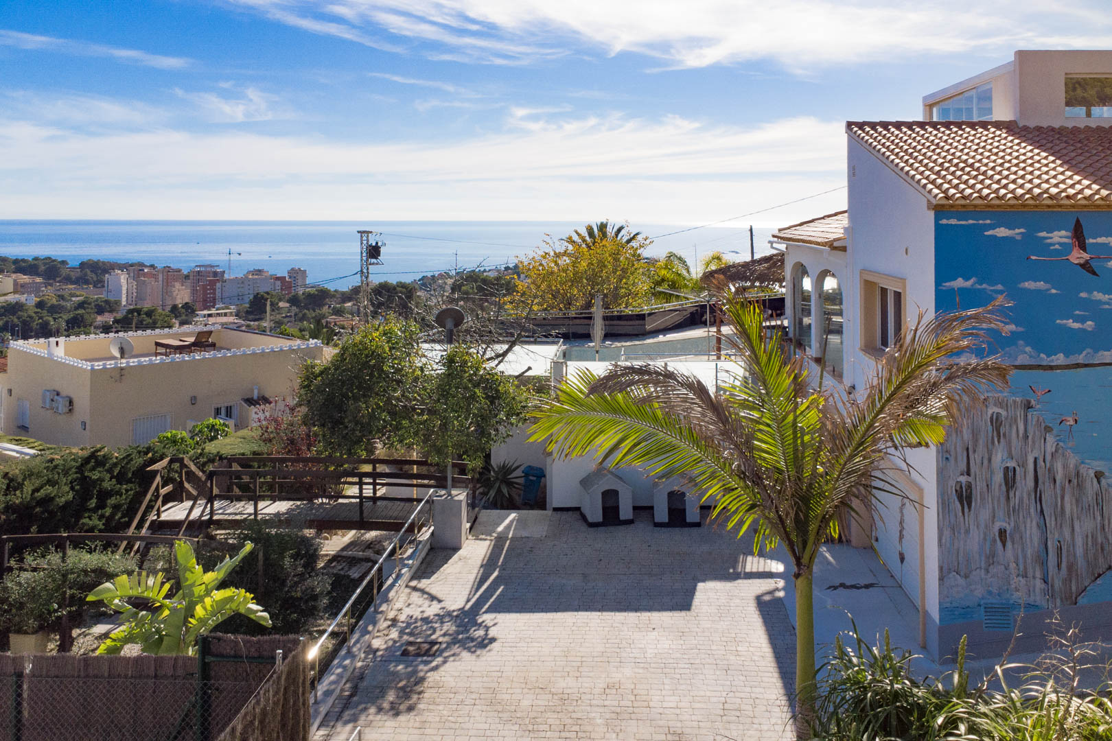 Vue extérieure avec entrée pavée, fresque murale de flamants roses et vue panoramique sur la mer Méditerranée et la silhouette de Calpe.