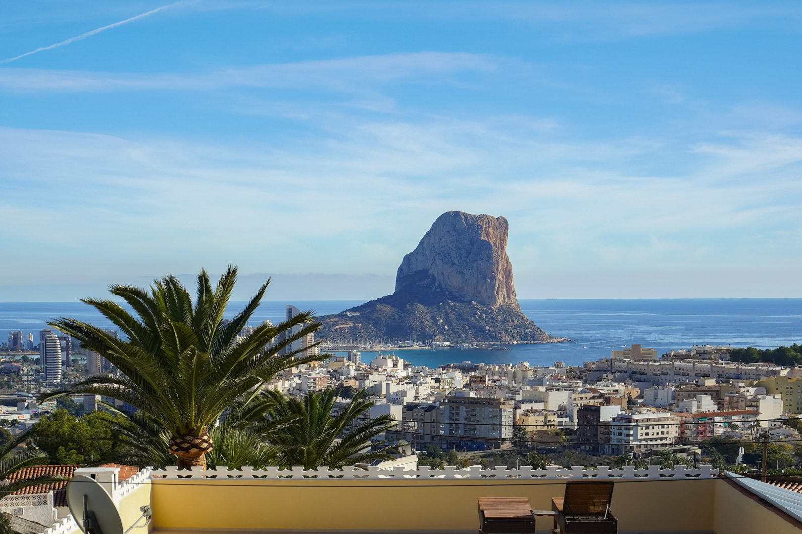Vue panoramique depuis la terrasse sur le Peñón de Ifach et la ville de Calpe, avec la mer Méditerranée en arrière-plan et des chaises longues en bois.