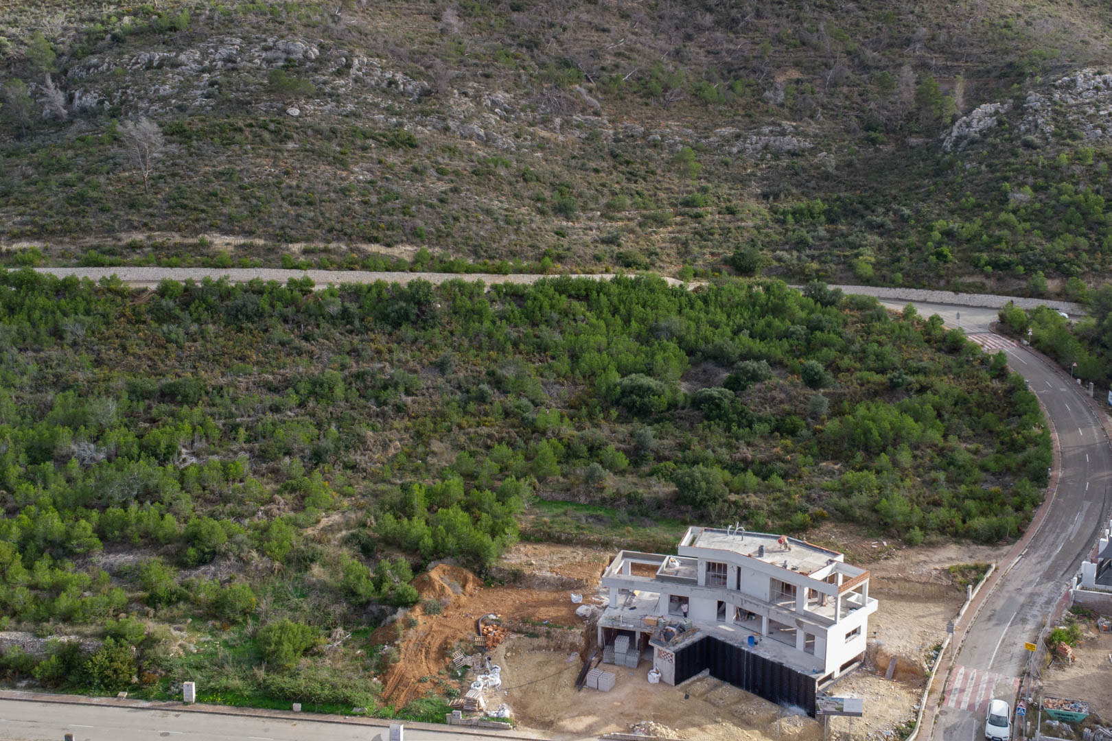 Vue aérienne d'une villa en cours de construction à Monte Olímpico, Jávea, entourée de collines verdoyantes et de routes d'accès pavées.