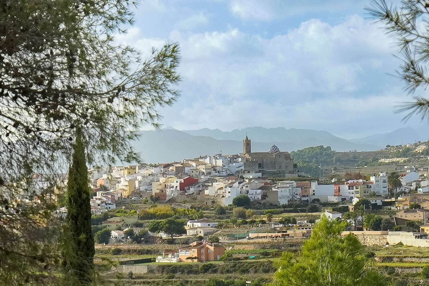 Vues panoramiques sur le centre historique de Benitachell et son église, encadrées par des pins avec la Sierra de Bernia en arrière-plan.