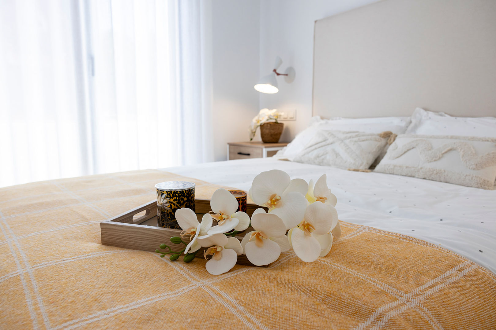 Chambre lumineuse avec tête de lit rembourrée, textiles blancs et moutarde, et plateau décoratif avec orchidées. La lumière naturelle traverse de grands voilages blancs.