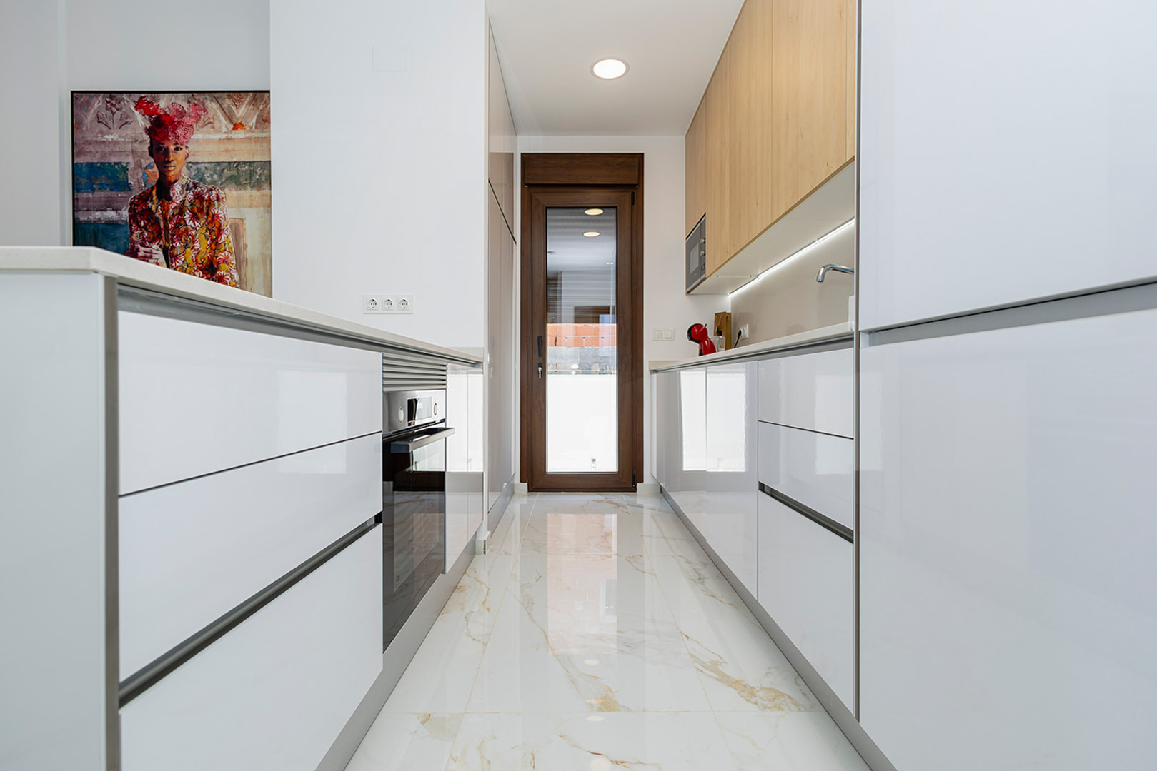Cuisine moderne avec placards blanc brillant, meubles hauts en bois et sol en grès cérame effet marbre. Équipée d'appareils intégrés et d'un accès direct vers l'extérieur.
