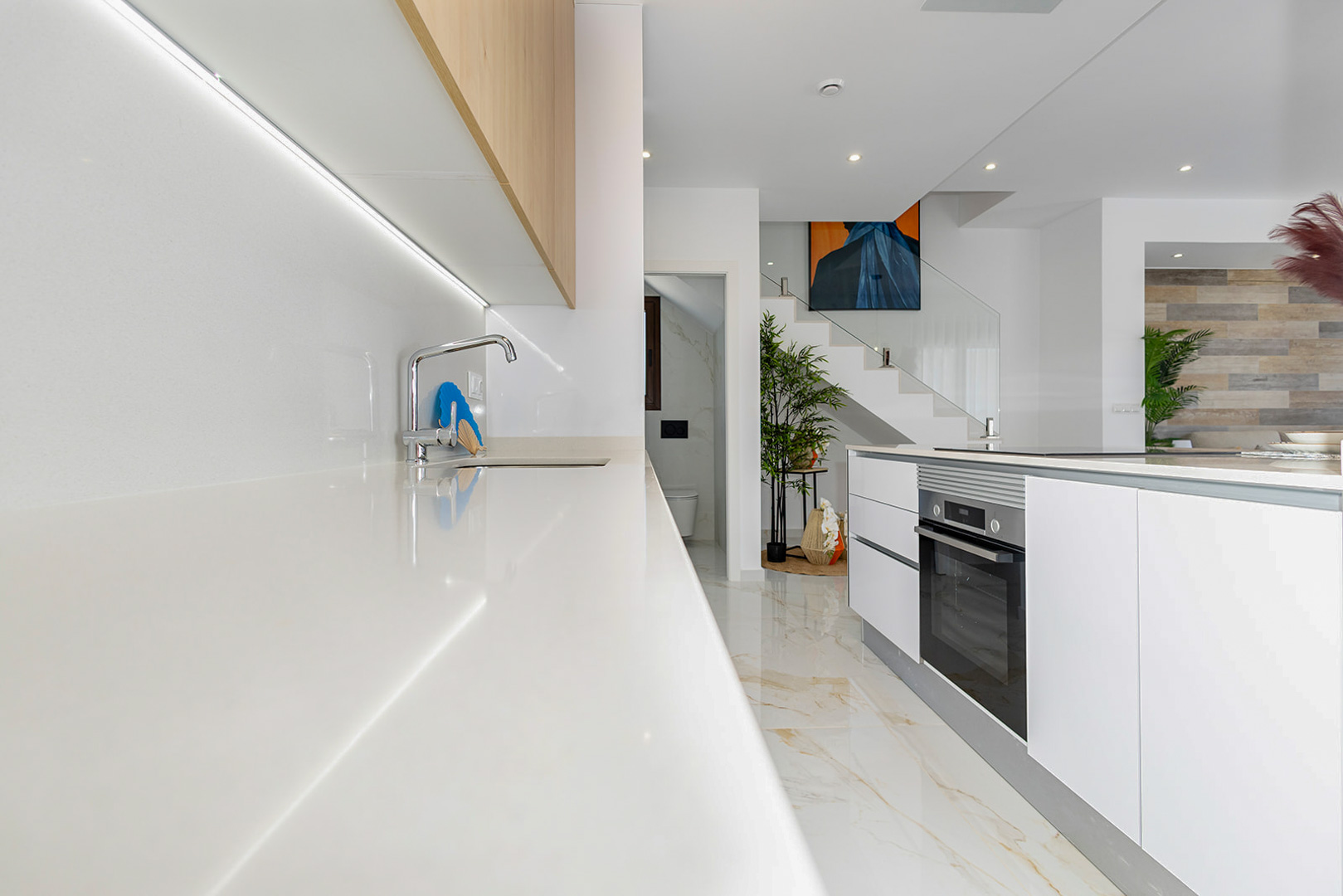 Cuisine moderne avec plan de travail blanc, éclairage LED sous les meubles en bois et îlot central. L'arrière-plan révèle un escalier avec garde-corps en verre.