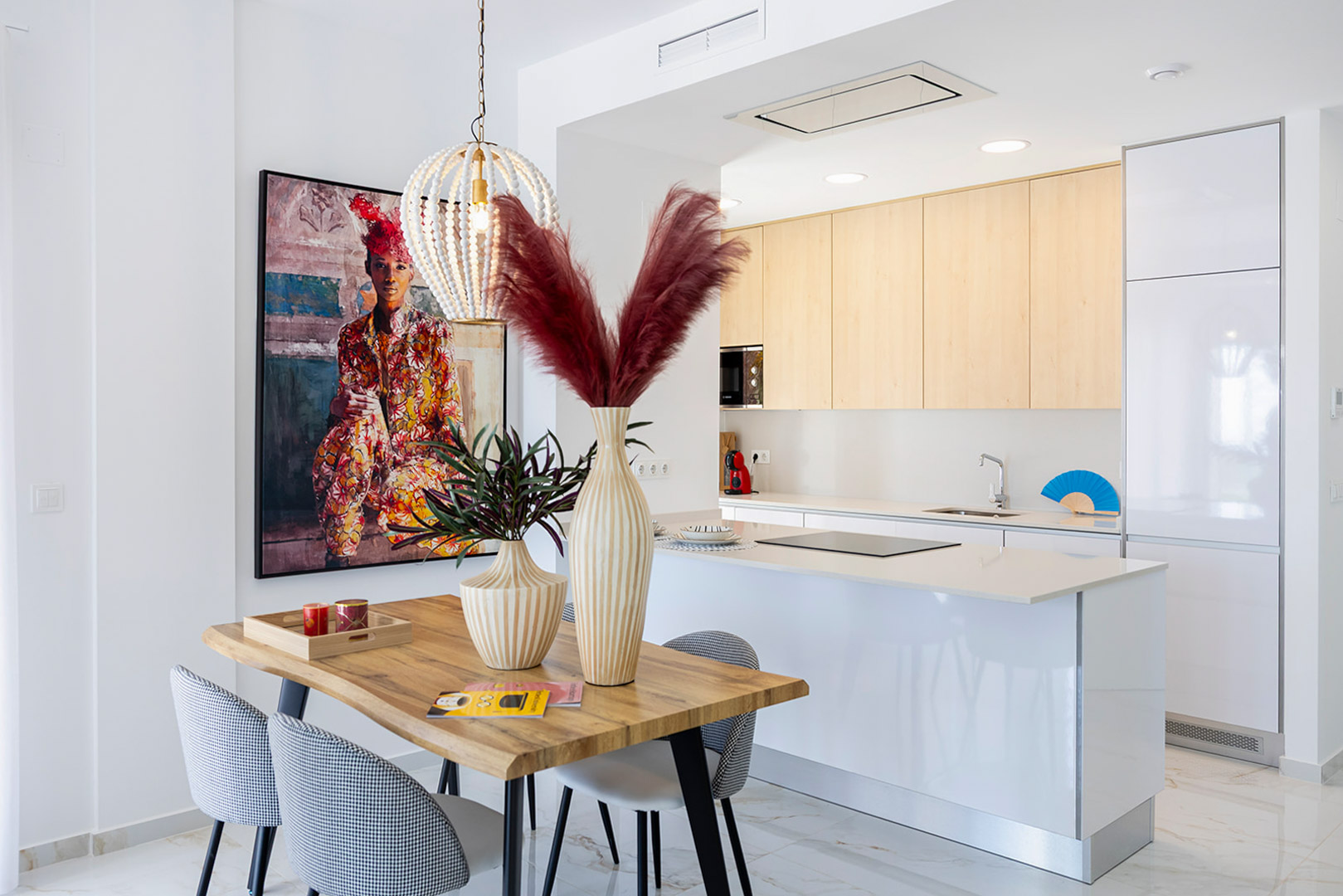 Cuisine ouverte avec îlot blanc et placards en bois clair, intégrée à un coin repas avec table en bois, chaises rembourrées et suspension décorative en perles.