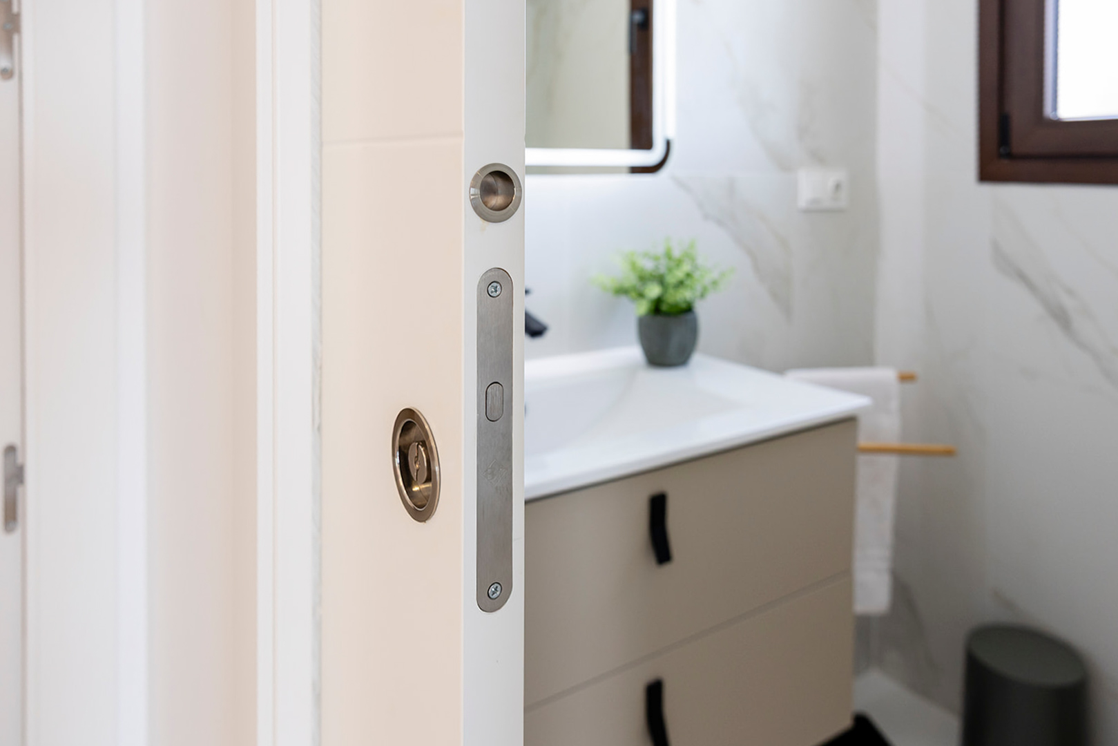 Détail d'une porte coulissante blanche avec poignée encastrée, ouvrant sur une salle de bain moderne avec carrelage effet marbre et meuble sous-vasque épuré.