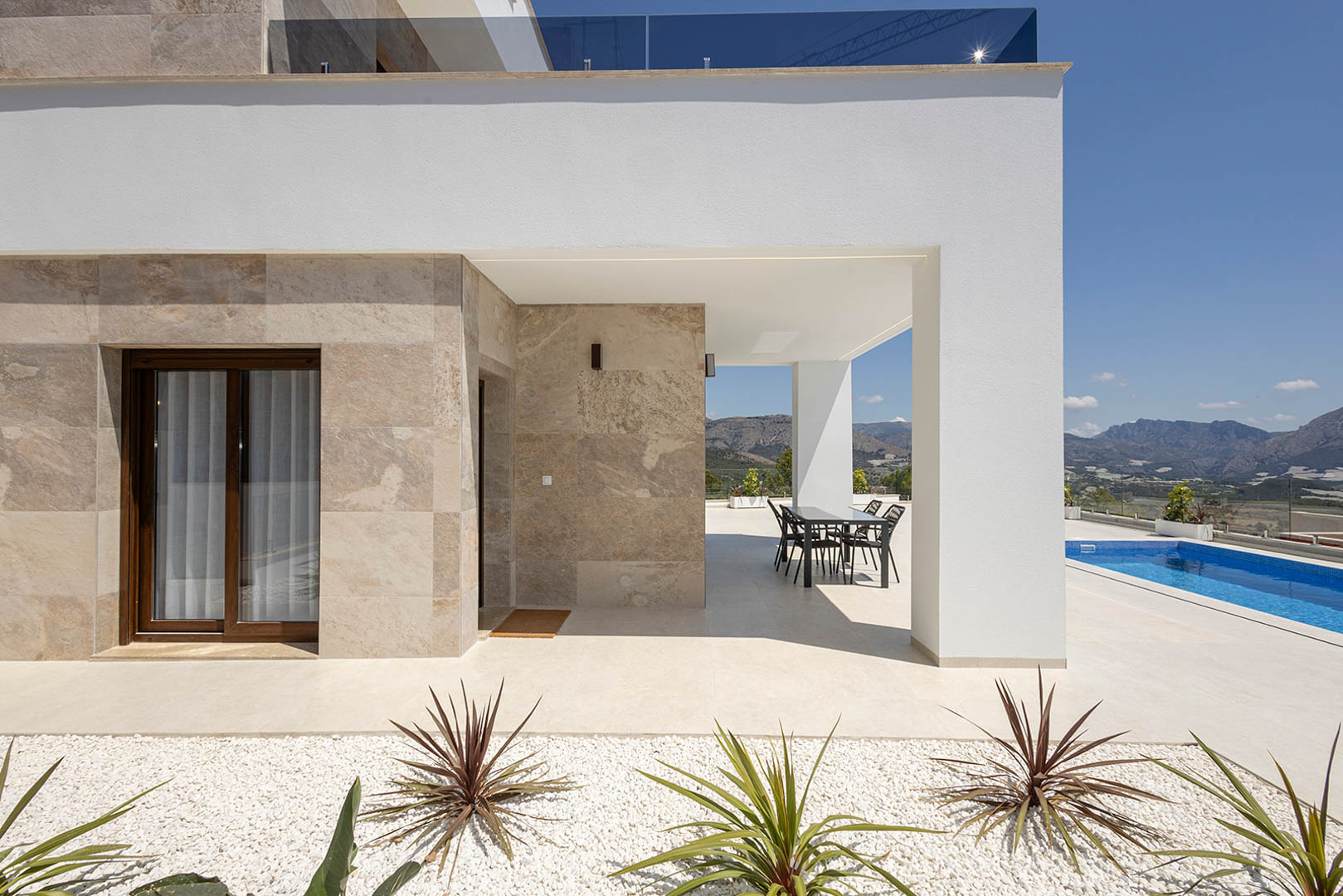 Extérieur de villa moderne avec parement en pierre naturelle, terrasse couverte, piscine privée et vue dégagée sur les montagnes environnantes.
