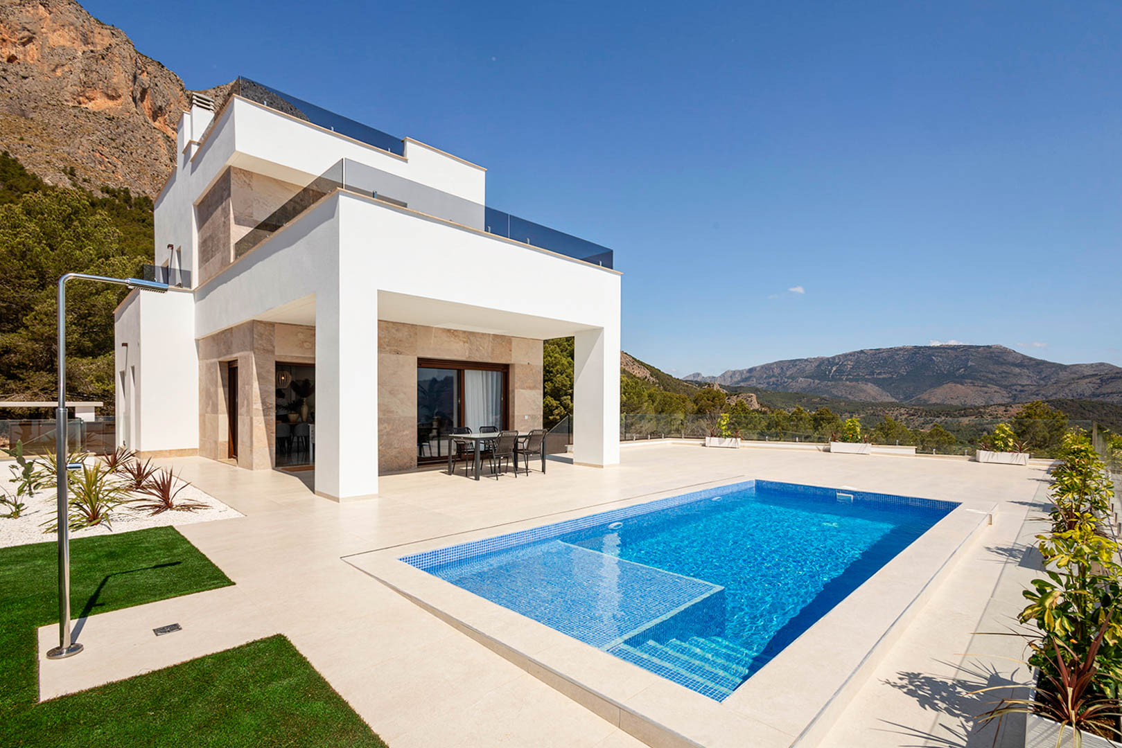 Extérieur d'une villa moderne avec façades blanches, parement en pierre, piscine privée, douche extérieure et terrasse offrant une vue sur les montagnes.
