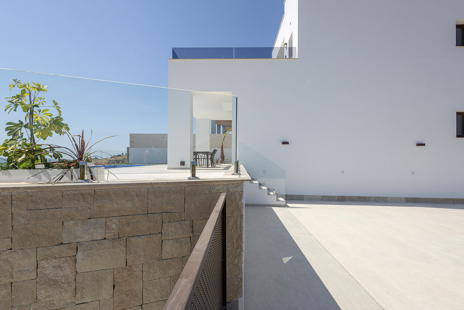 Façade blanche aux lignes épurées avec mur en pierre naturelle, garde-corps en verre et espace extérieur pavé menant à la terrasse supérieure.