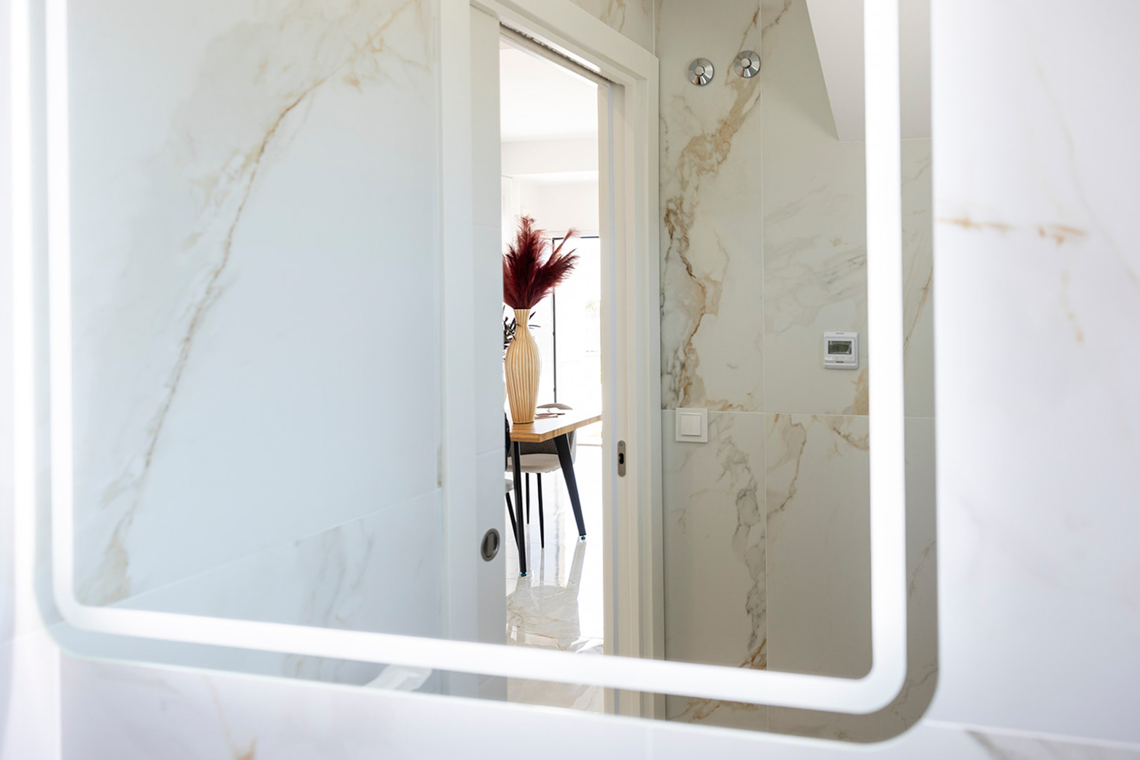 Salle de bain avec carrelage effet marbre et miroir rétroéclairé par LED, offrant une vue sur la salle à manger à travers une porte blanche.