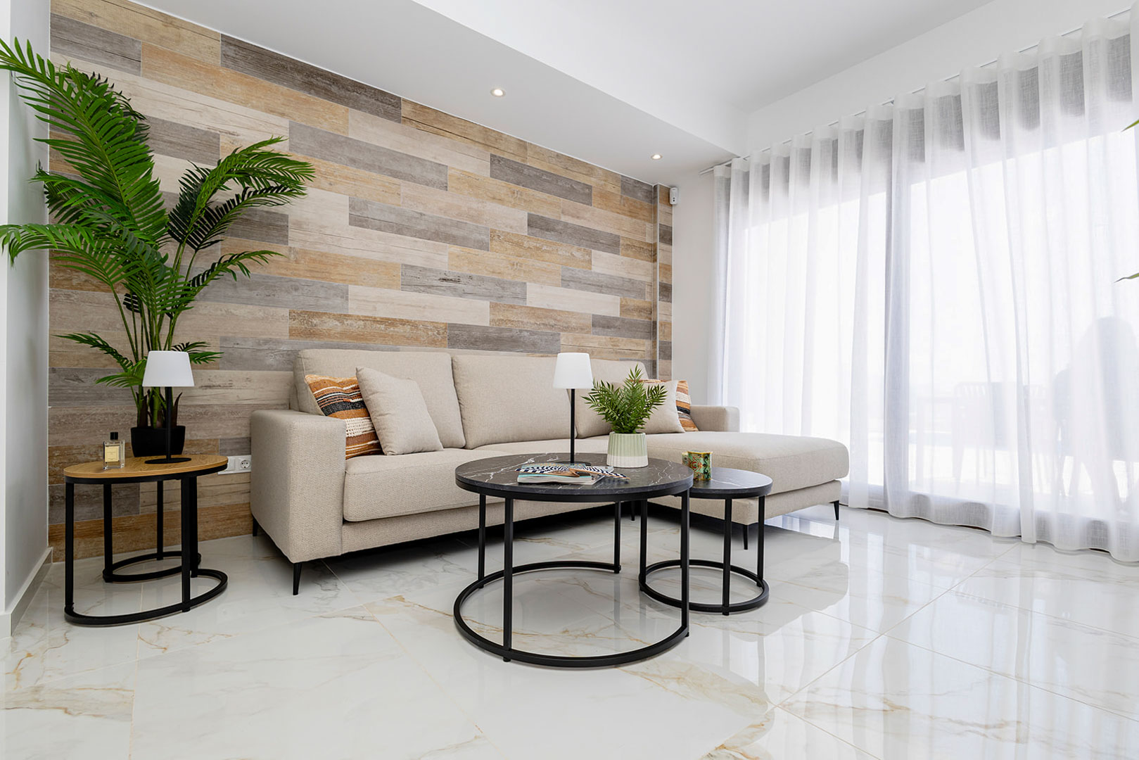 Salon avec mur d'accent en lattes de bois, sol en grès cérame effet marbre et canapé beige bordé de grandes baies vitrées aux rideaux voilages.