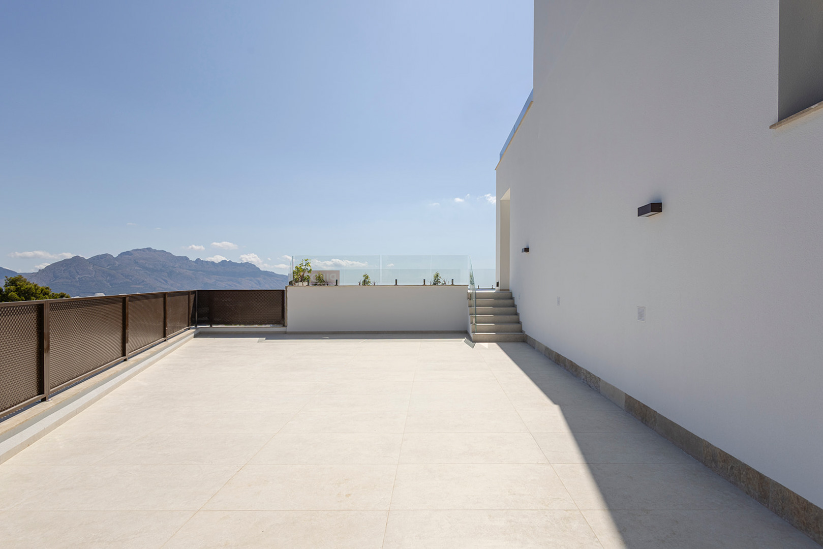Terrasse supérieure avec carrelage clair, garde-corps en verre et vue dégagée sur les montagnes. Éclairage extérieur moderne intégré à la façade blanche.