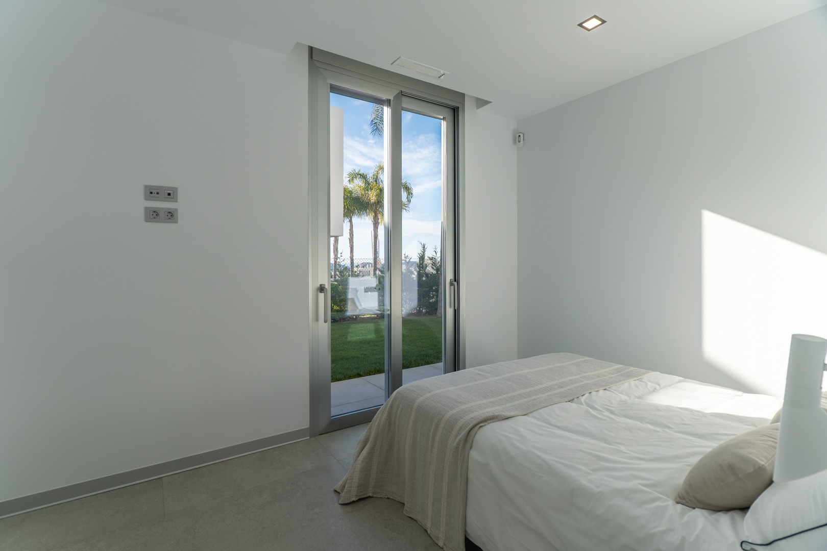 Chambre lumineuse avec accès direct au jardin par une baie vitrée, sols en grès cérame gris et murs blancs au design épuré.