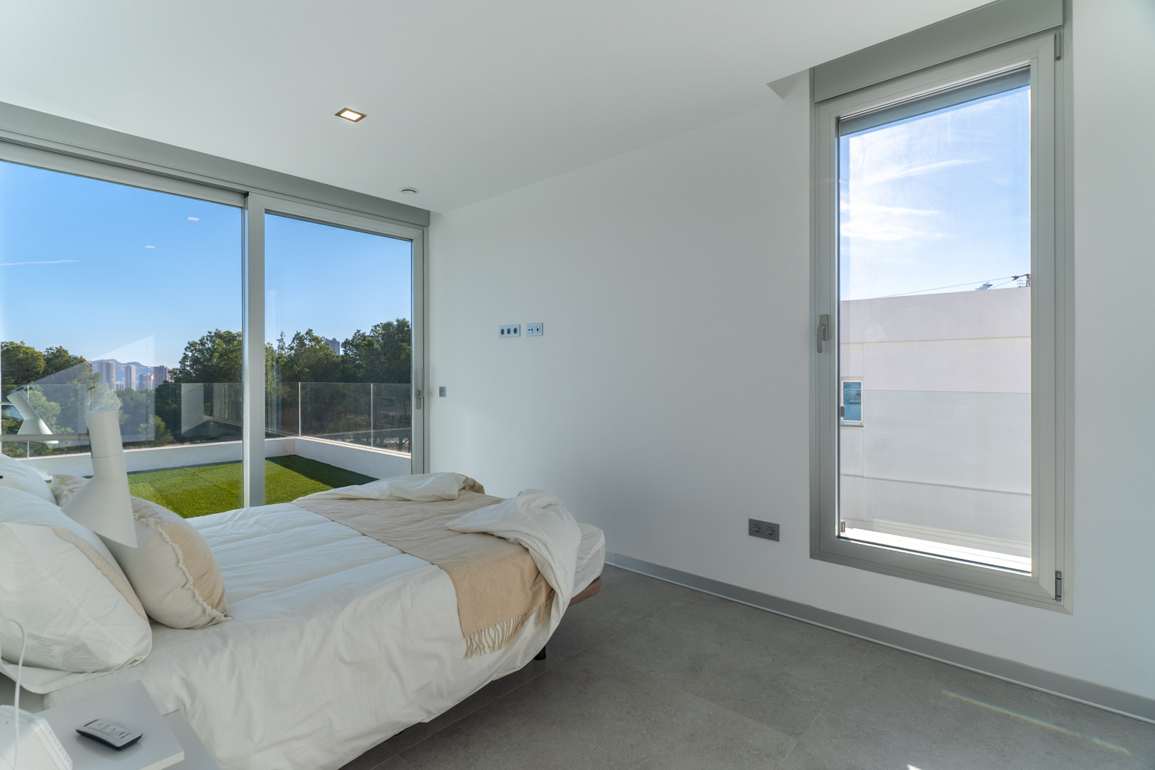 Chambre lumineuse avec sol en grès cérame gris, grandes baies vitrées en aluminium et accès direct à une terrasse privée avec gazon synthétique.