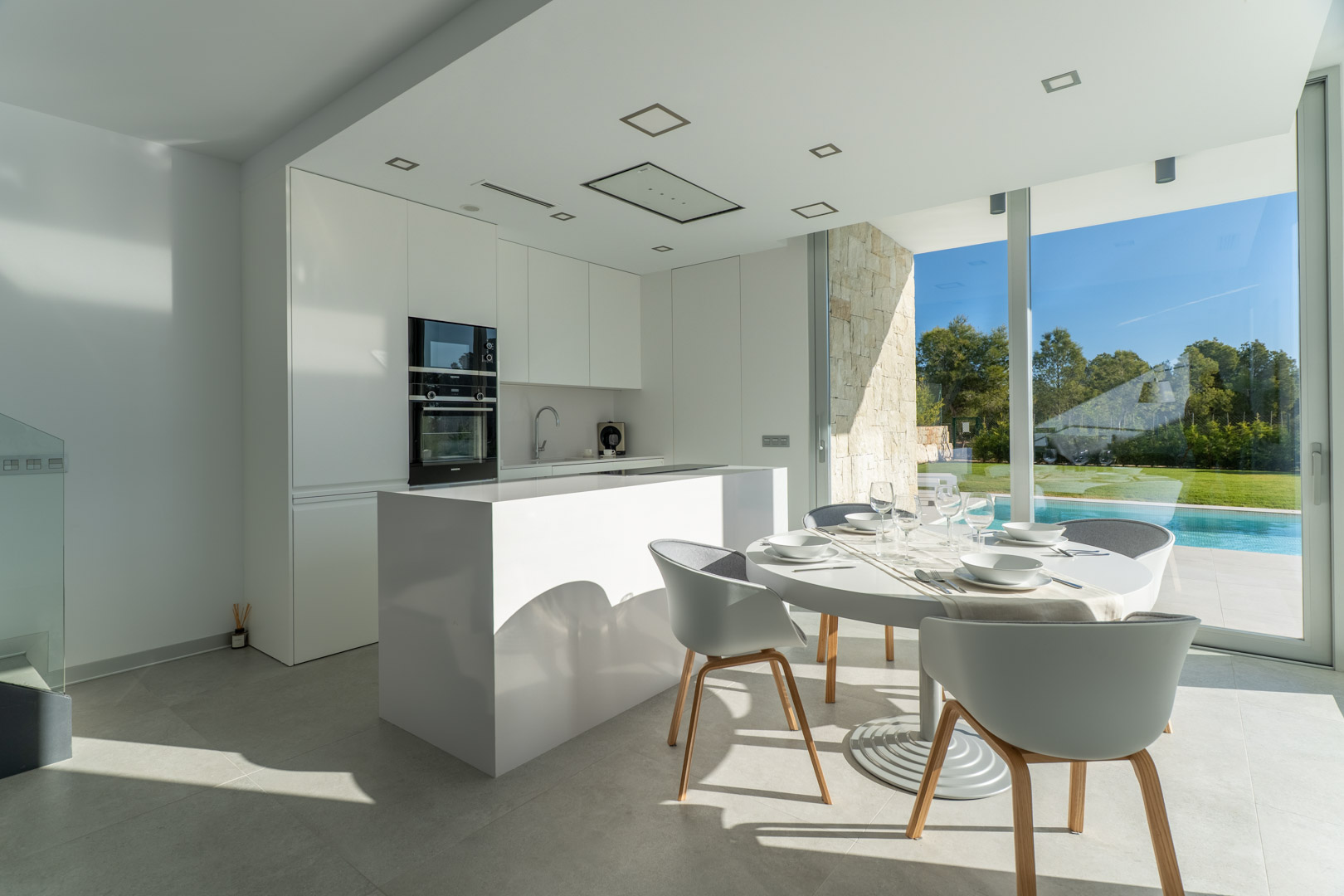 Cuisine américaine minimaliste blanche avec îlot central, ouverte sur un espace repas lumineux offrant une vue directe sur la piscine et le jardin.