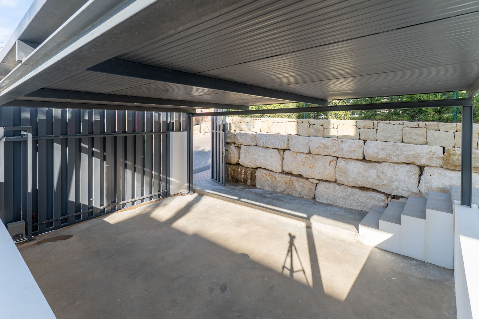 Espace de stationnement couvert avec structure métallique, sol en béton et mur de soutènement en pierre naturelle, incluant un accès par portail.