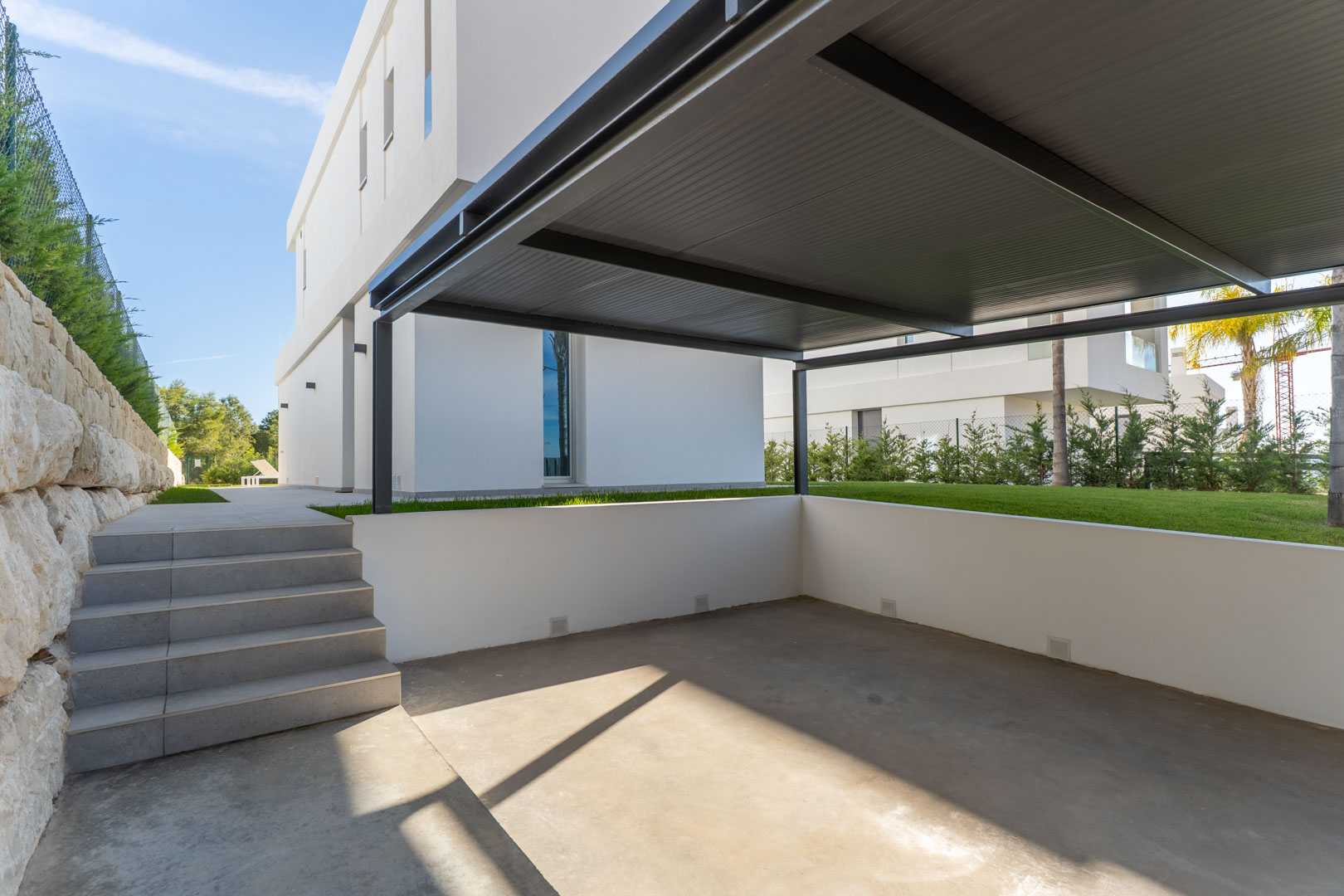 Espace de stationnement privé avec abri métallique, sol en béton et escaliers en grès menant au jardin et à la façade de la villa.