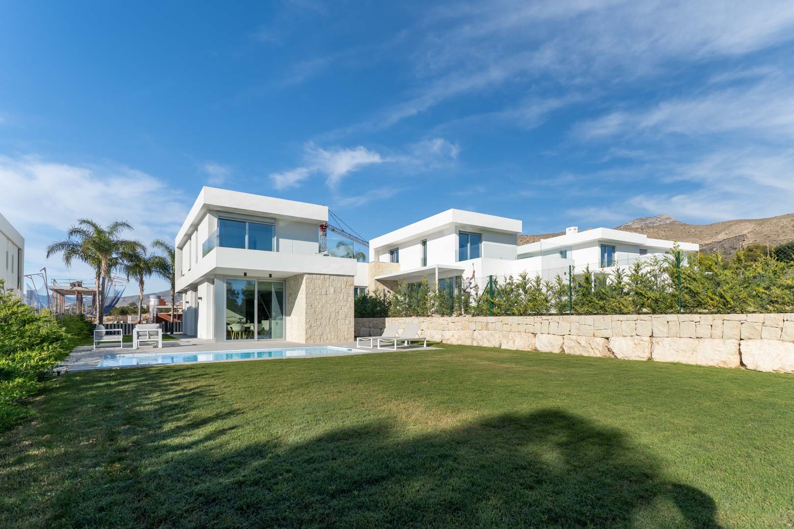 Extérieur de villa moderne avec façade blanche, détails en pierre, piscine privée, vaste jardin de pelouse et vue sur les montagnes environnantes.