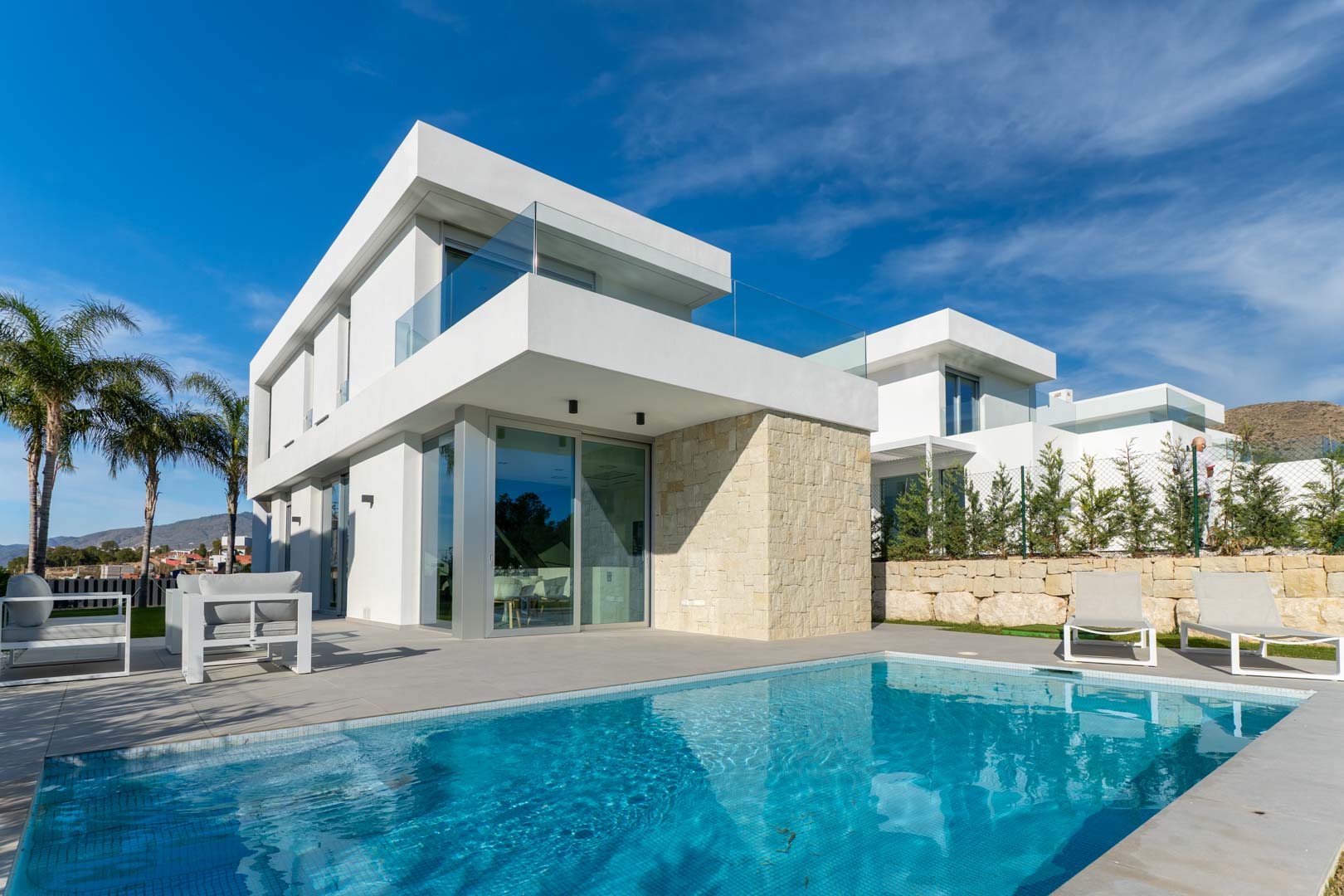 Extérieur de villa moderne avec piscine privée, façade blanche épurée, mur en pierre naturelle et terrasses avec garde-corps en verre.