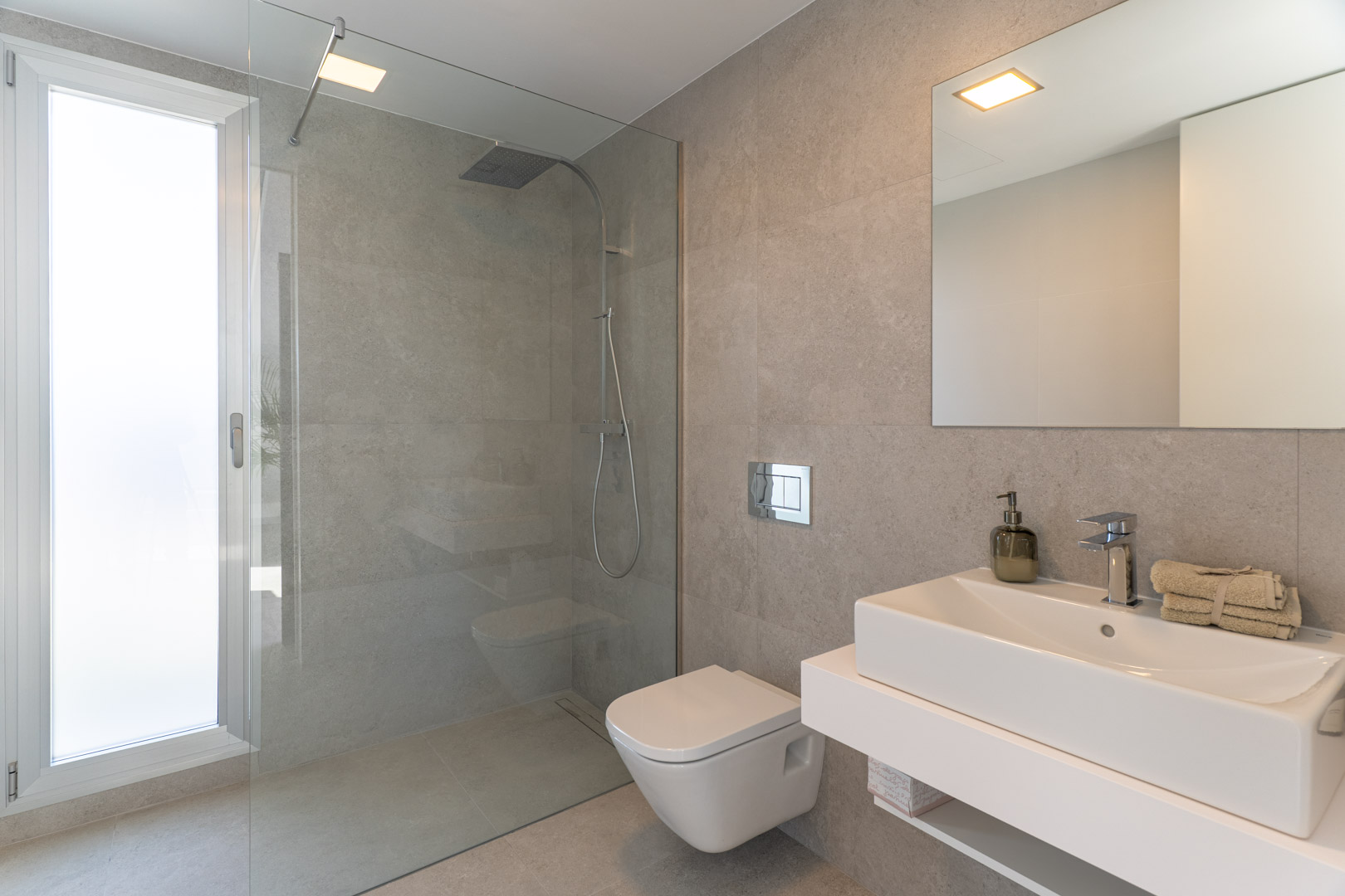 Salle de bains moderne avec carrelage gris, douche à l'italienne vitrée, WC suspendu et meuble vasque blanc suspendu sous un grand miroir.