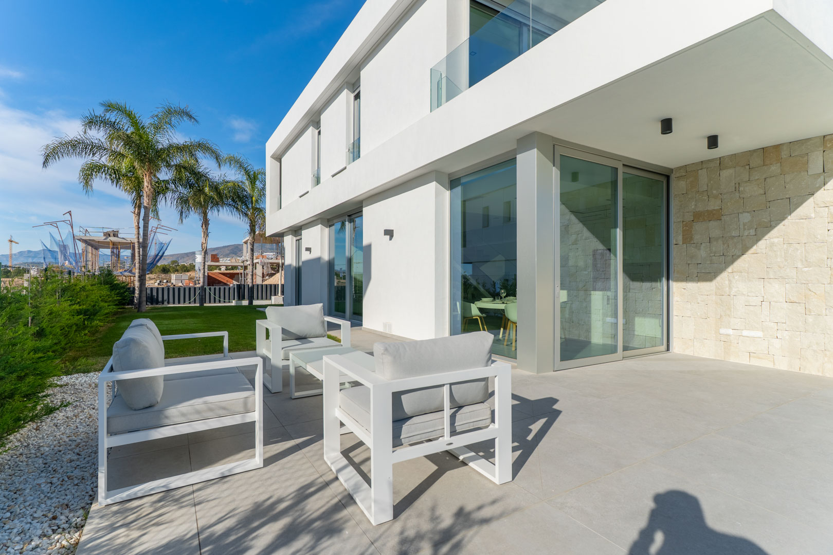 Terrasse extérieure avec mobilier minimaliste blanc, sol en grès cérame et façade alliant enduit blanc et mur en pierre naturelle texturée.