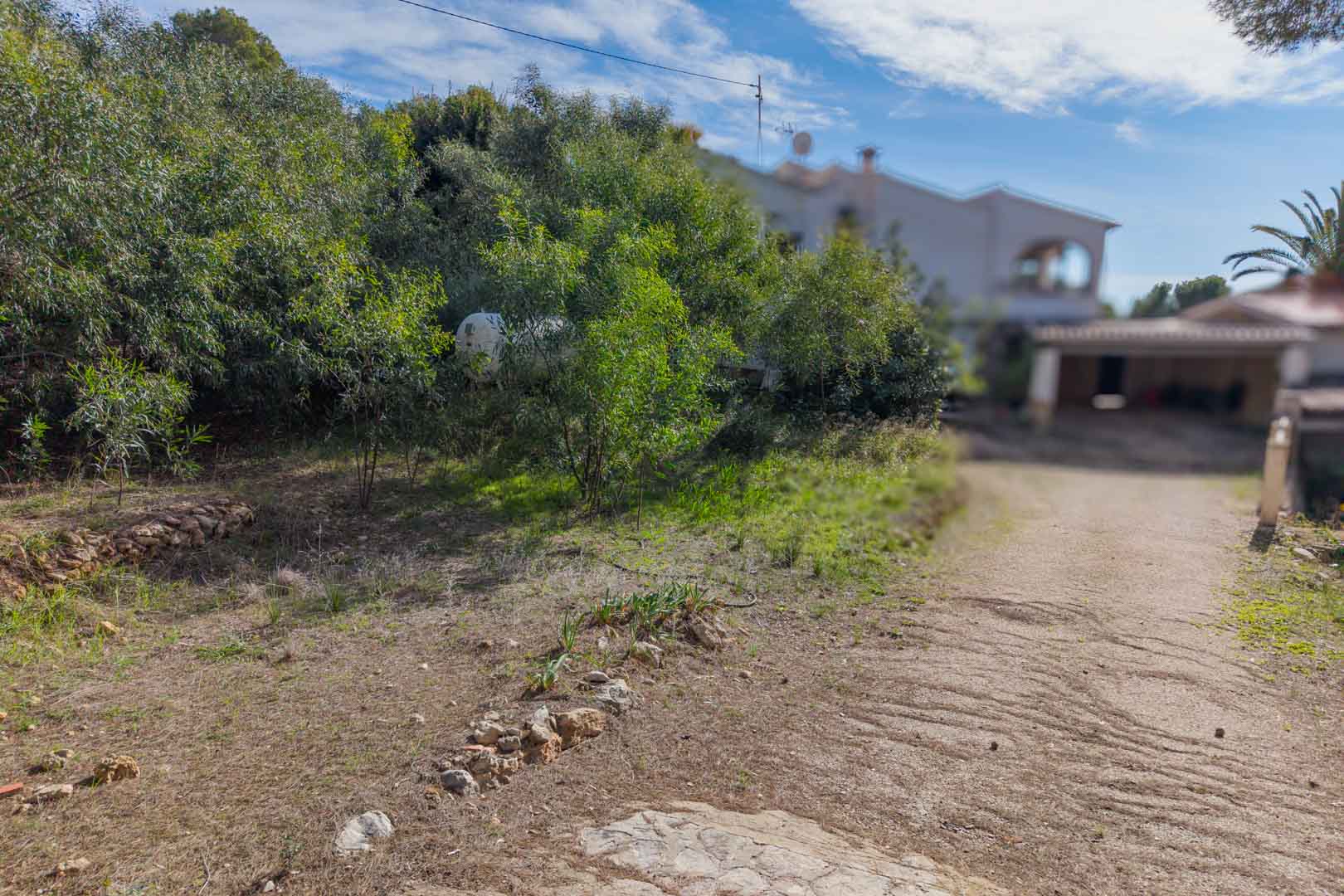 Chemin d'accès au terrain urbain avec bordures en pierre naturelle et végétation méditerranéenne, incluant une vue sur la villa voisine.