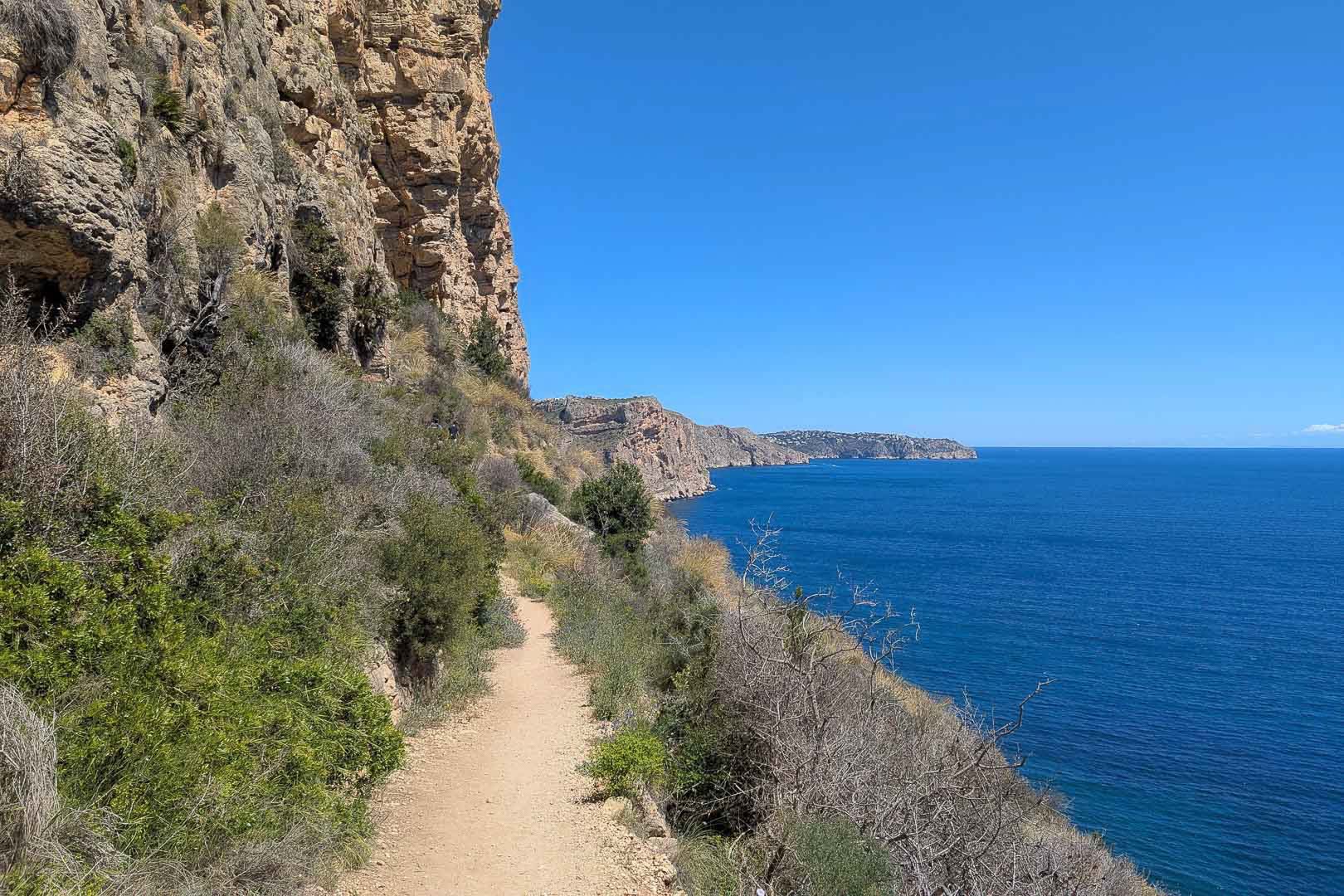 Sentier côtier en terre bordant des falaises calcaires avec vue panoramique sur la mer Méditerranée et la végétation locale par temps clair.